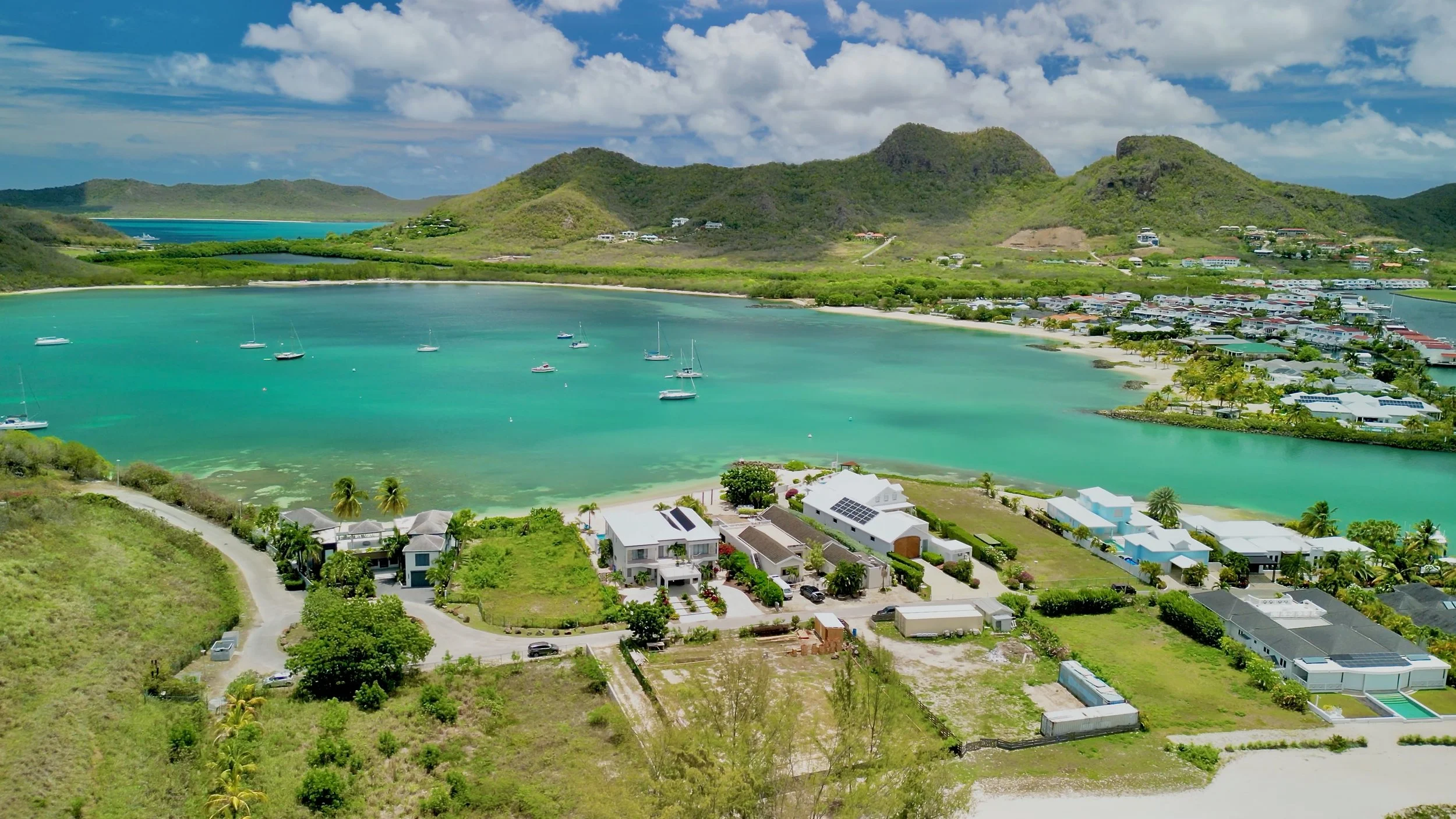 An aerial view of a tropical island featuring turquoise water, sailboats, houses, lush green hills, and a partly cloudy sky.