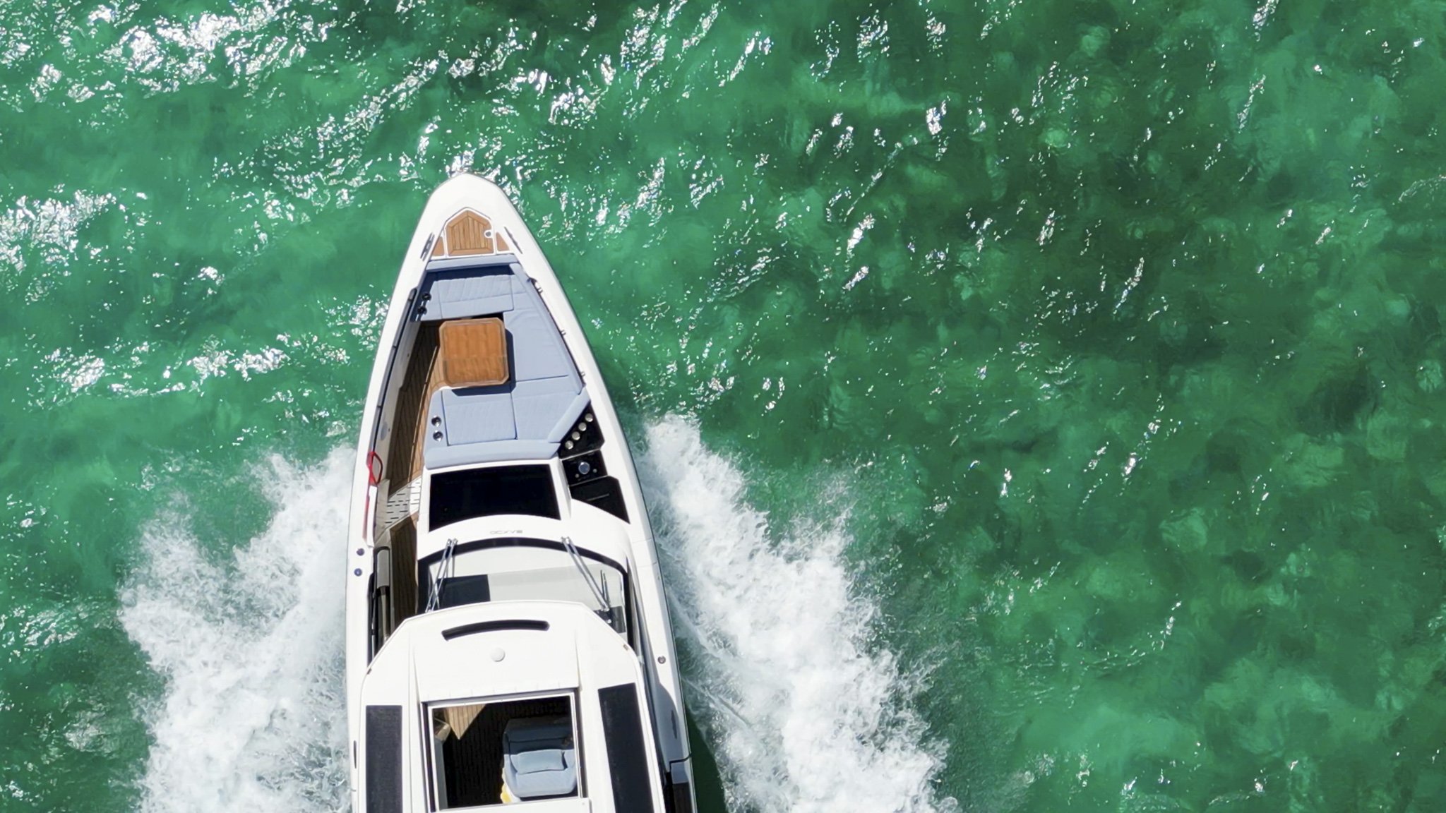 Top-down view of a white motor yacht moving through clear turquoise water, creating a wake behind it, with wooden deck and black accents visible.
