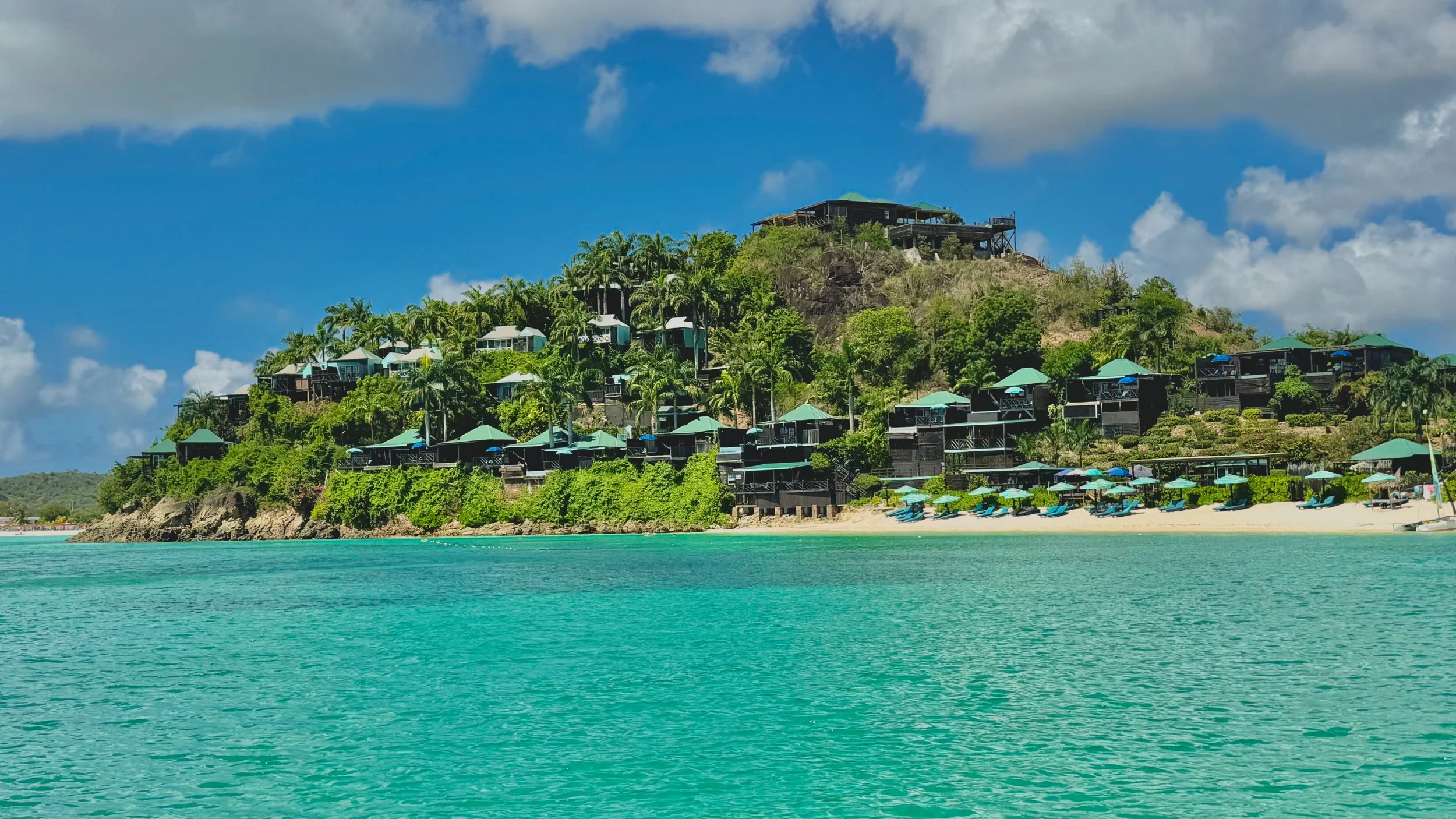A tropical beach with turquoise water, a sandy shoreline with blue lounge chairs and umbrellas, and a hillside with green trees and modern houses with green roofs.