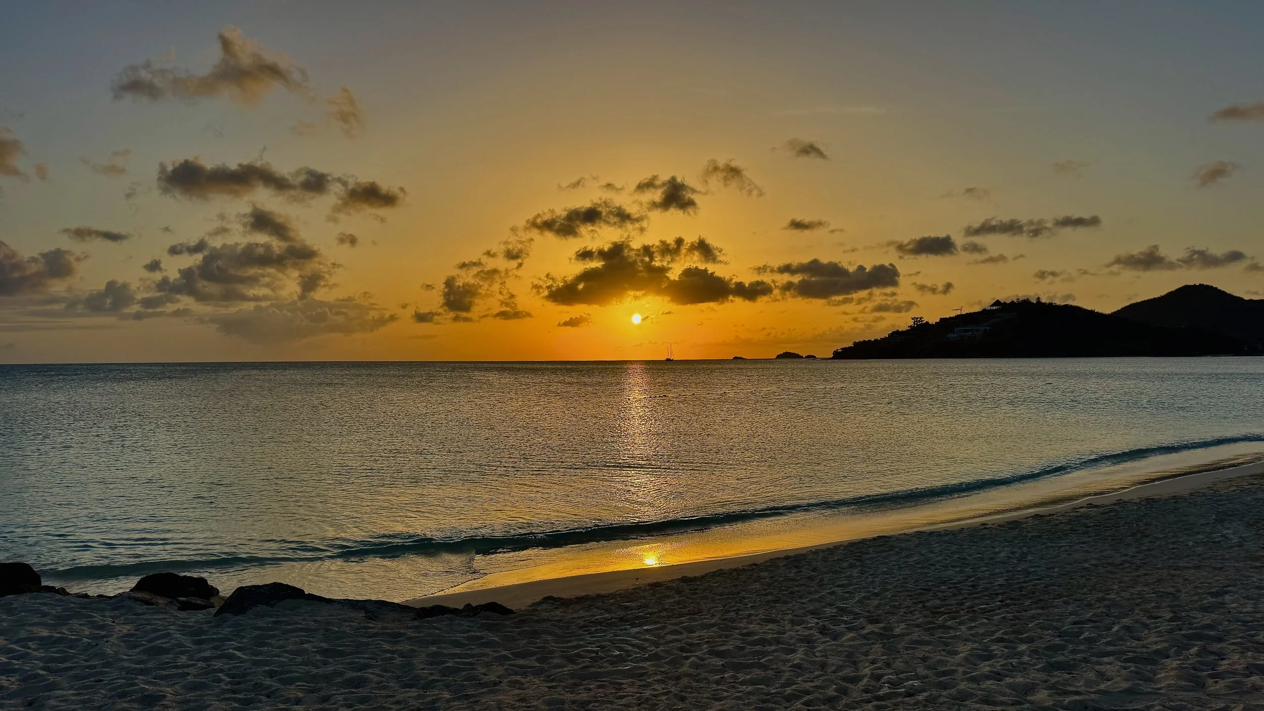 Sunset over the ocean with a partly cloudy sky, distant sailboat, and a hill with houses on the right, viewed from a sandy beach with rocks in the foreground.