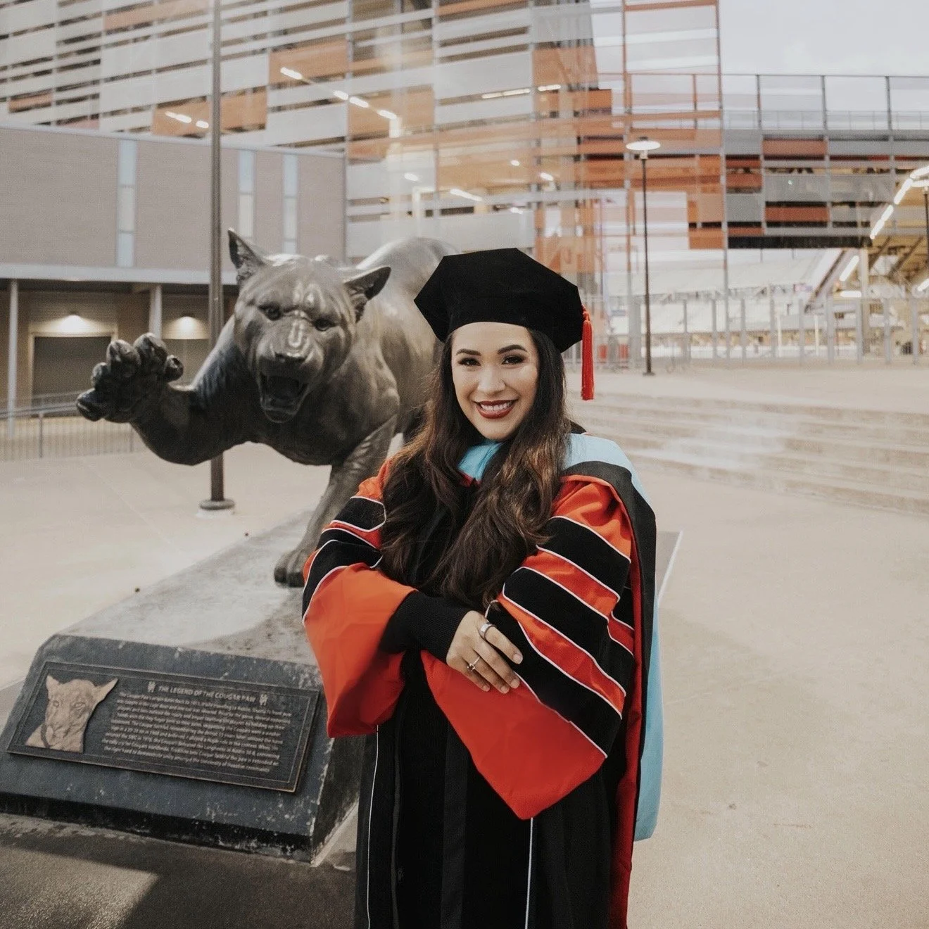 A young woman in graduation attire standing in front of a bronze statue of a lion. She is smiling, wearing a black cap and gown with orange and black accents, and a light blue hood. There is an informational plaque at the base of the statue.