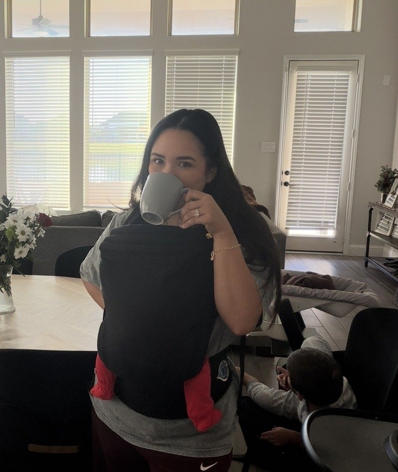 Woman with long dark hair holding a mug, looking into a mirror, in a well-lit living room with large windows, a dining table with a flower arrangement, and a child sitting in a chair nearby.