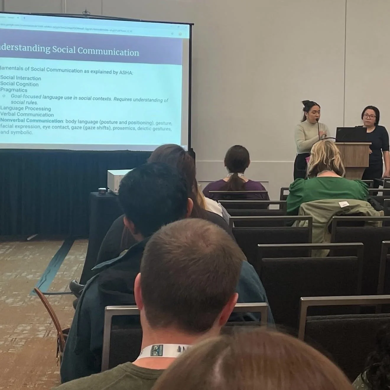 A presentation on social communication fundamentals is being given in a conference room. Two women are presenting at a podium, while an audience observes and takes notes.