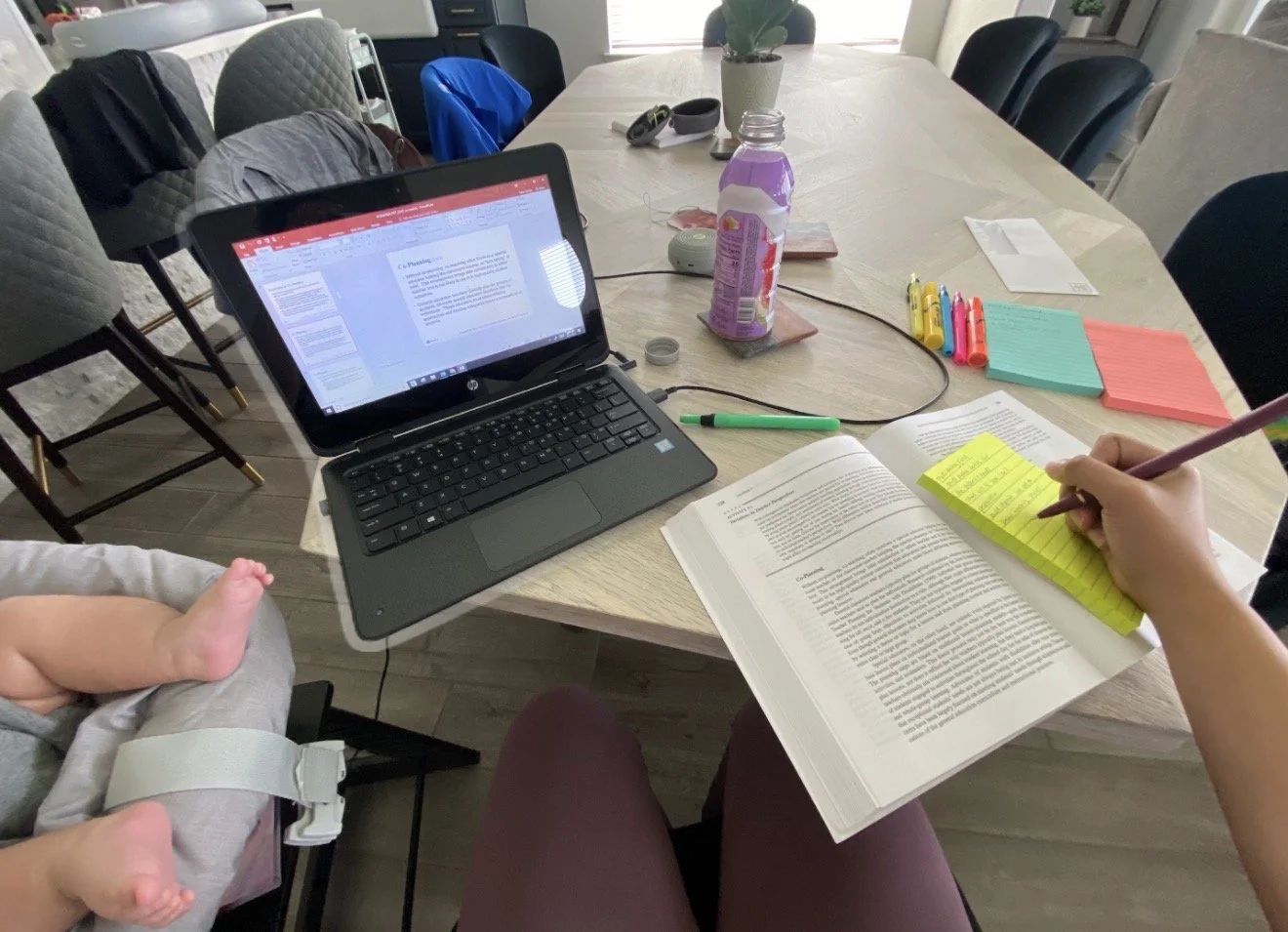 A person working at a dining table with a laptop, open book, highlighters, sticky notes, and a child in a baby car seat nearby.