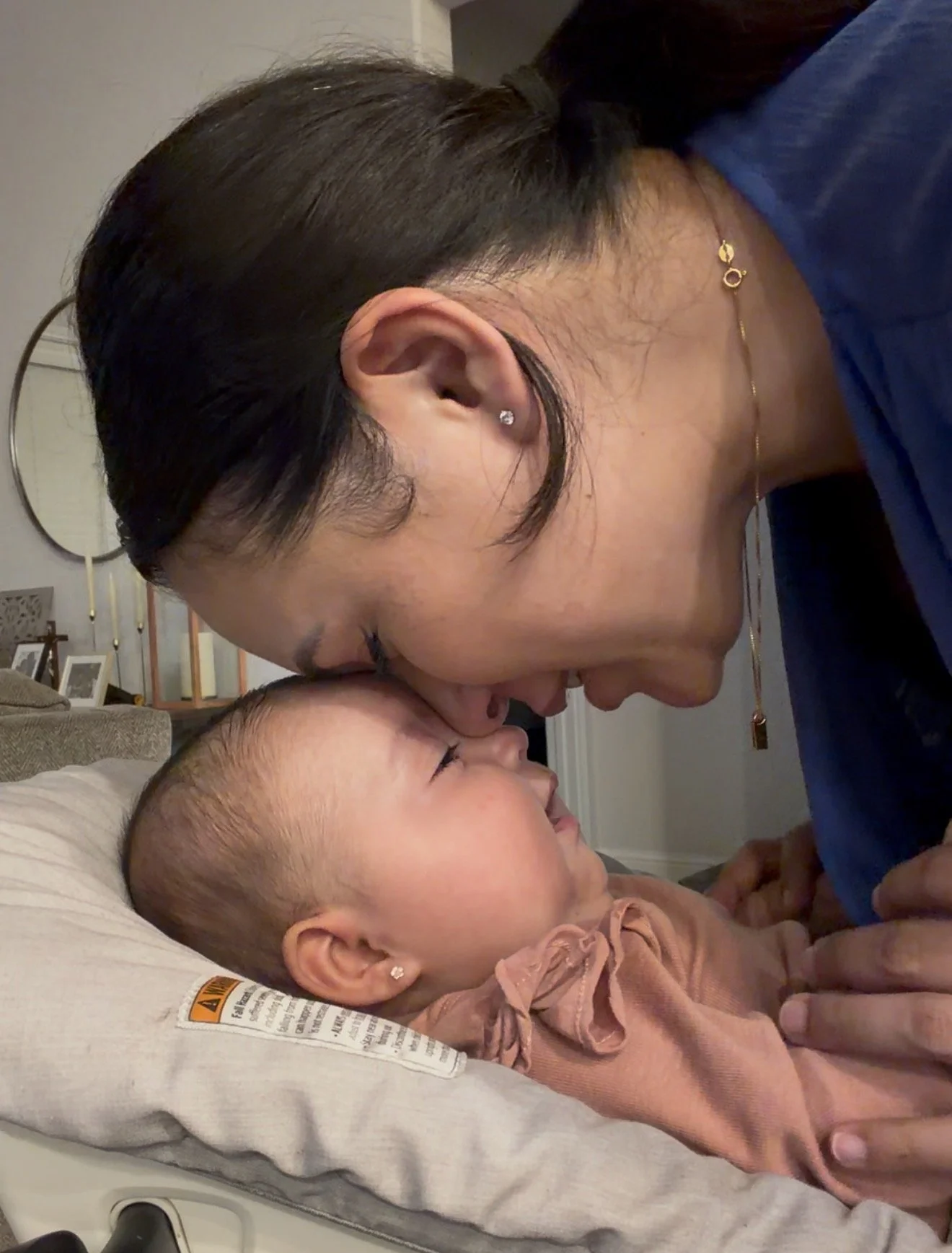 A woman and a young girl are sharing a close moment indoors. The woman is leaning over the girl with her forehead touching the girl’s forehead, both with eyes closed, smiling. The girl is lying on a bed with a beige pillow, wearing a brown top, and the woman is wearing a blue top and gold jewelry.