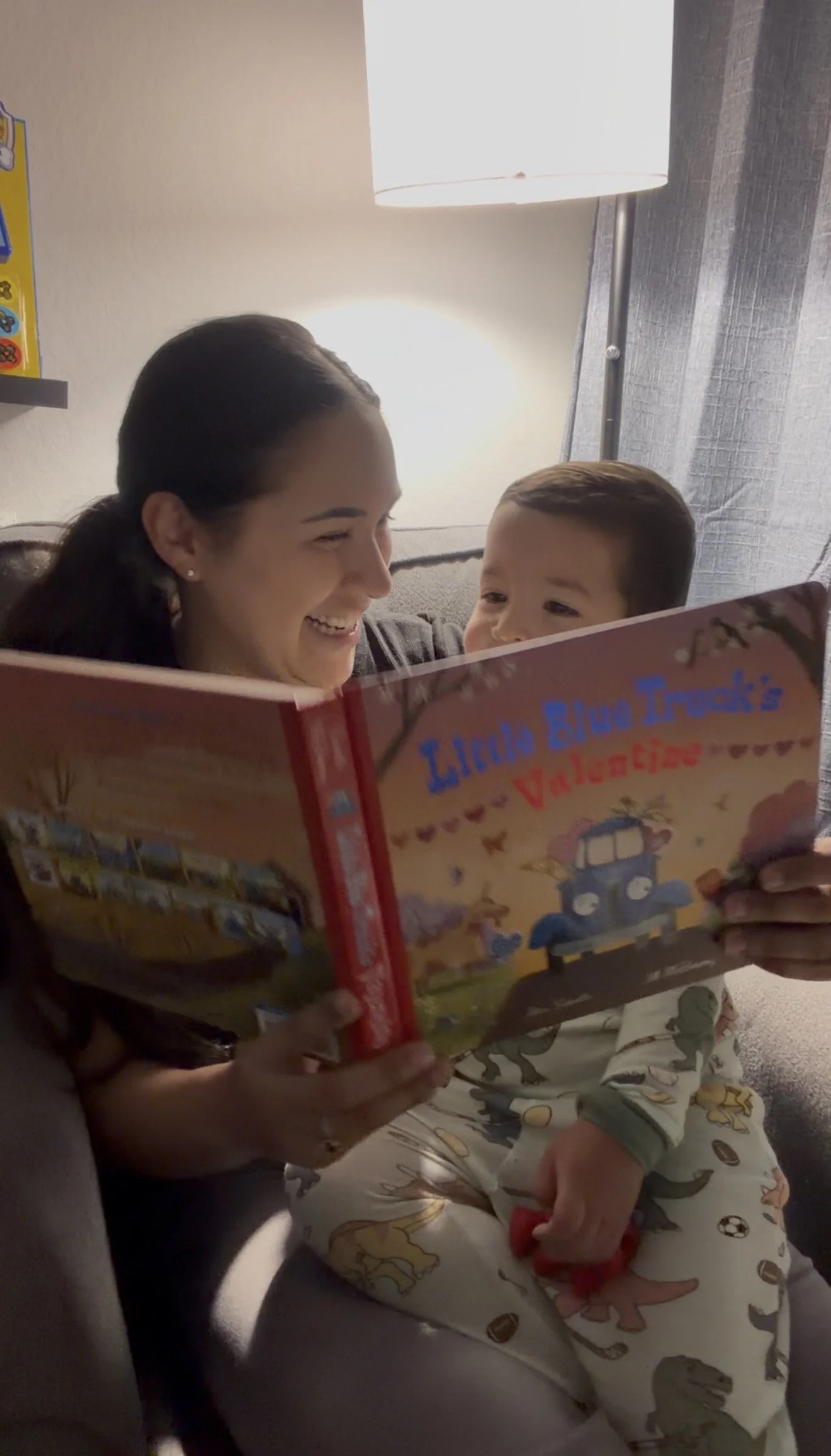 A woman and a young boy are sitting together on a couch, looking at a book titled 'Lucia Blu Truck's Valentine's.' The woman is smiling and has dark hair, while the boy has light brown hair and is wearing pajamas with dinosaur prints. They are near a lamp and a gray curtain.