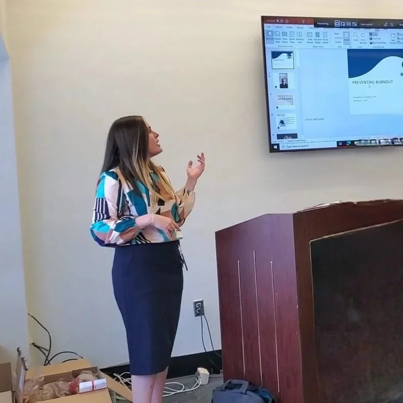 C.P. Handler giving a presentation in a conference room, standing next to a large screen displaying a slide titled 'Preventing Burnout'.
