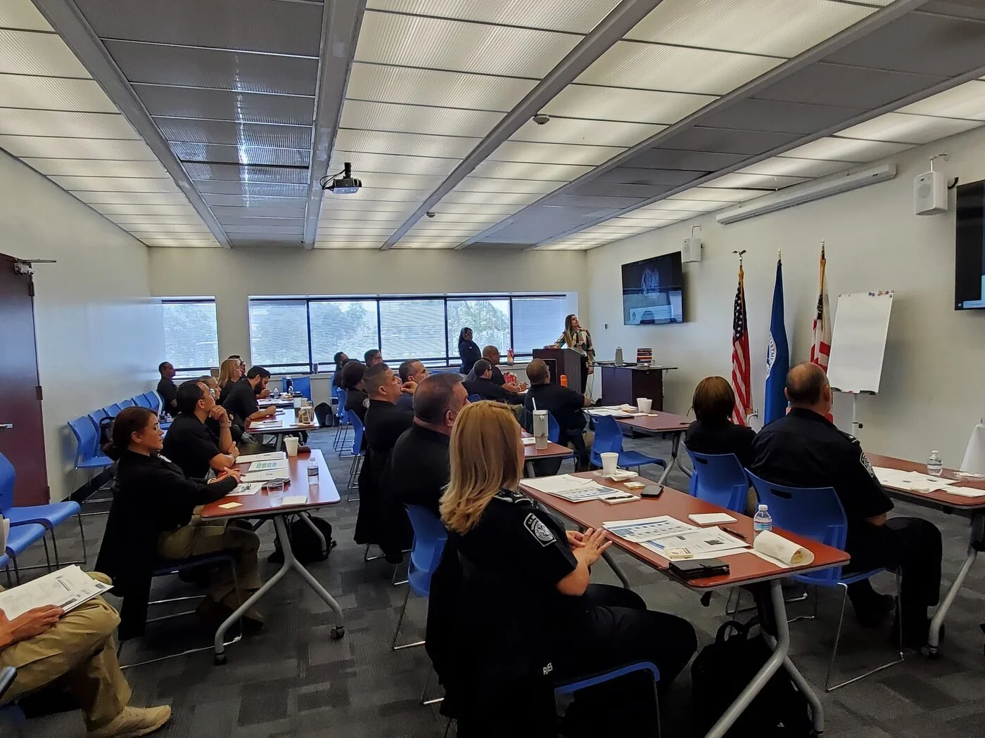 A classroom or conference room with badges of attendees, some in uniform. A woman at the podium appears to be giving a presentation, with seated attendees listening. Flags and screens are visible on the wall.