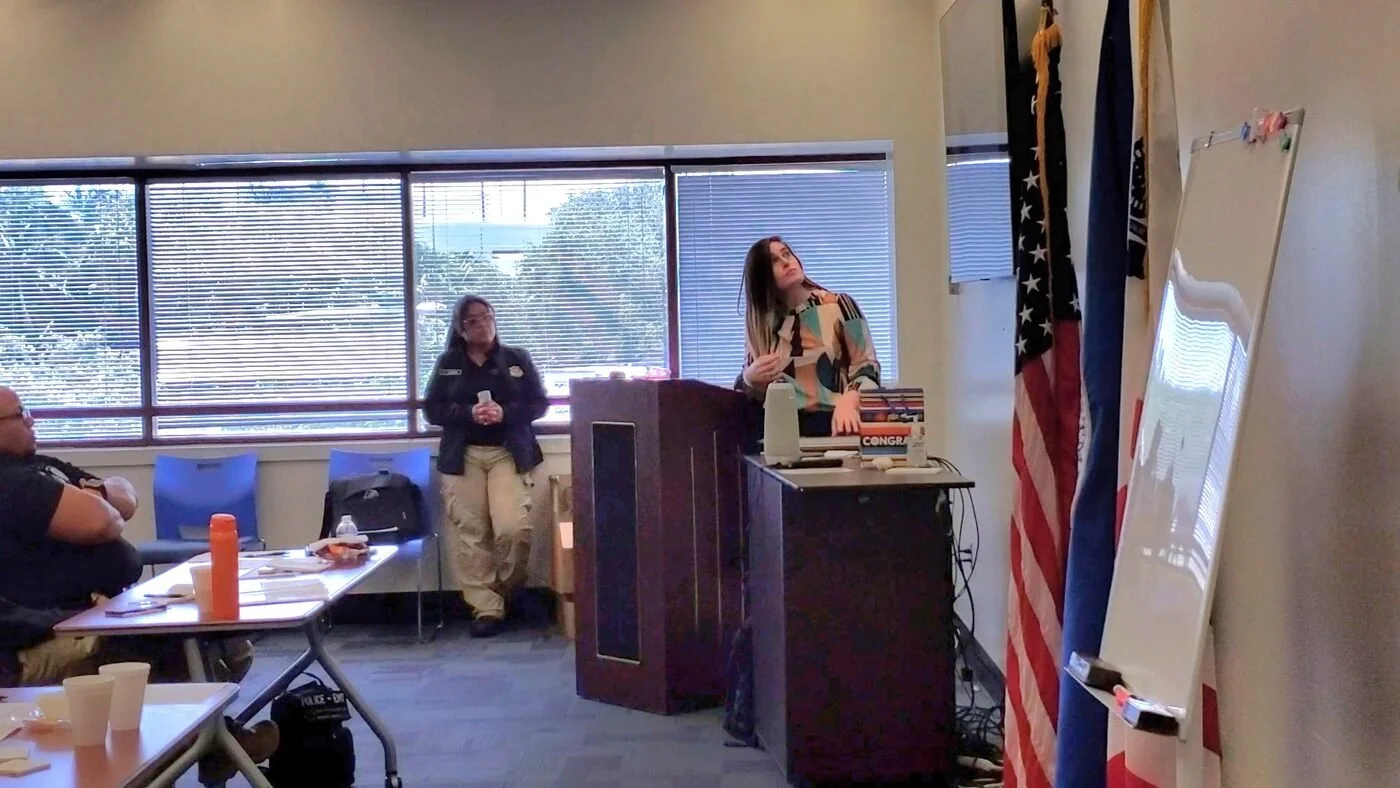 C.P. Handler standing at a podium giving a presentation in a room with several people seated and listening, with flags and a whiteboard nearby, and large windows in the background.