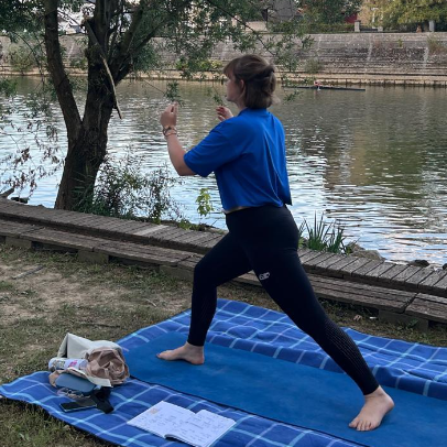 Femme faisant du yoga en position de guerrier au bord d'une rivière, sur un tapis d'exercice avec un sac et un journal à côté.