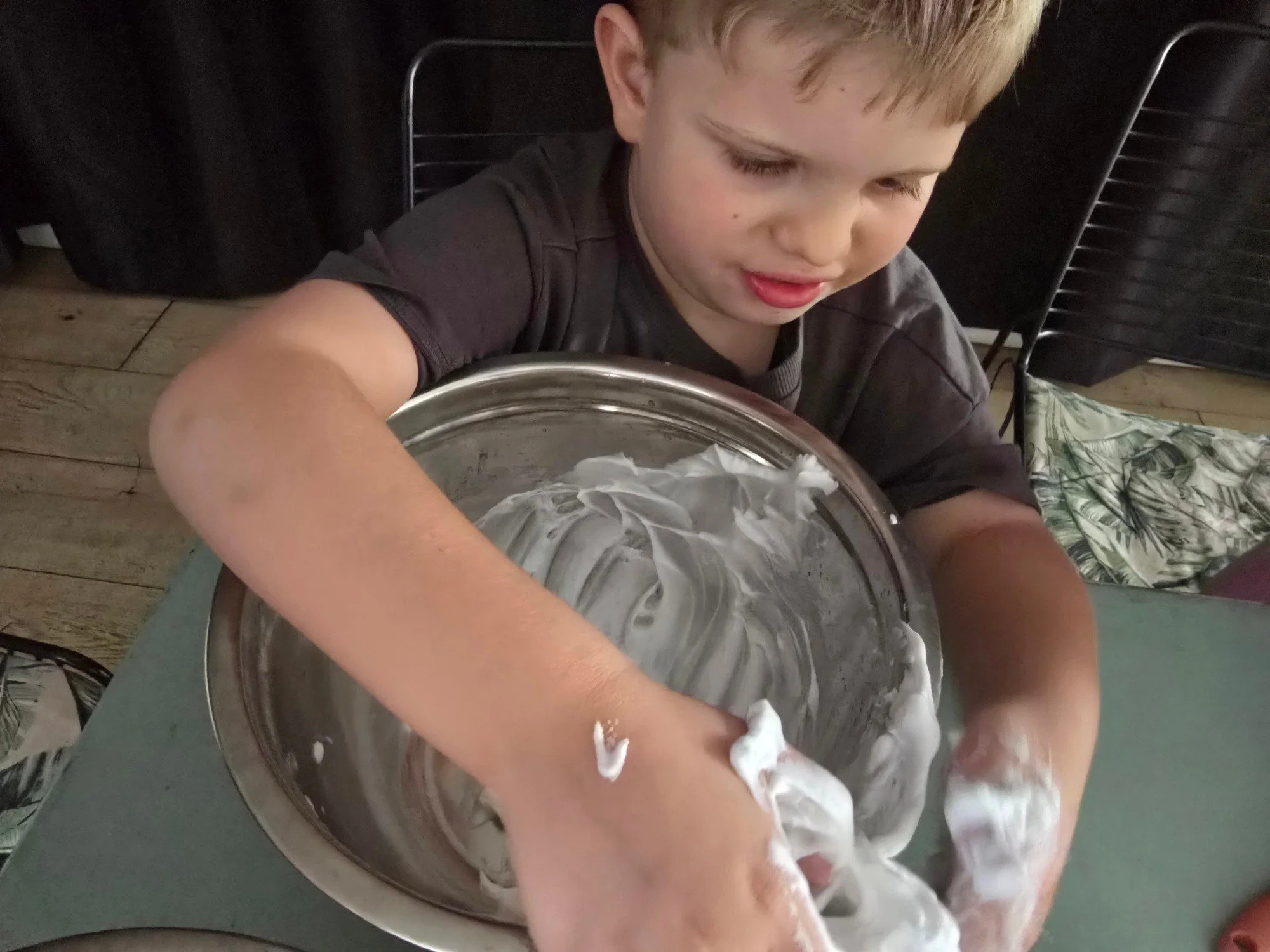 A young boy with blonde hair is engaging in a hands-on activity, mixing or kneading a fluffy white substance, possibly whipped cream or shaving foam, in a large metal bowl. The setting appears to be indoors with dark chairs and a wooden floor visible in the background.