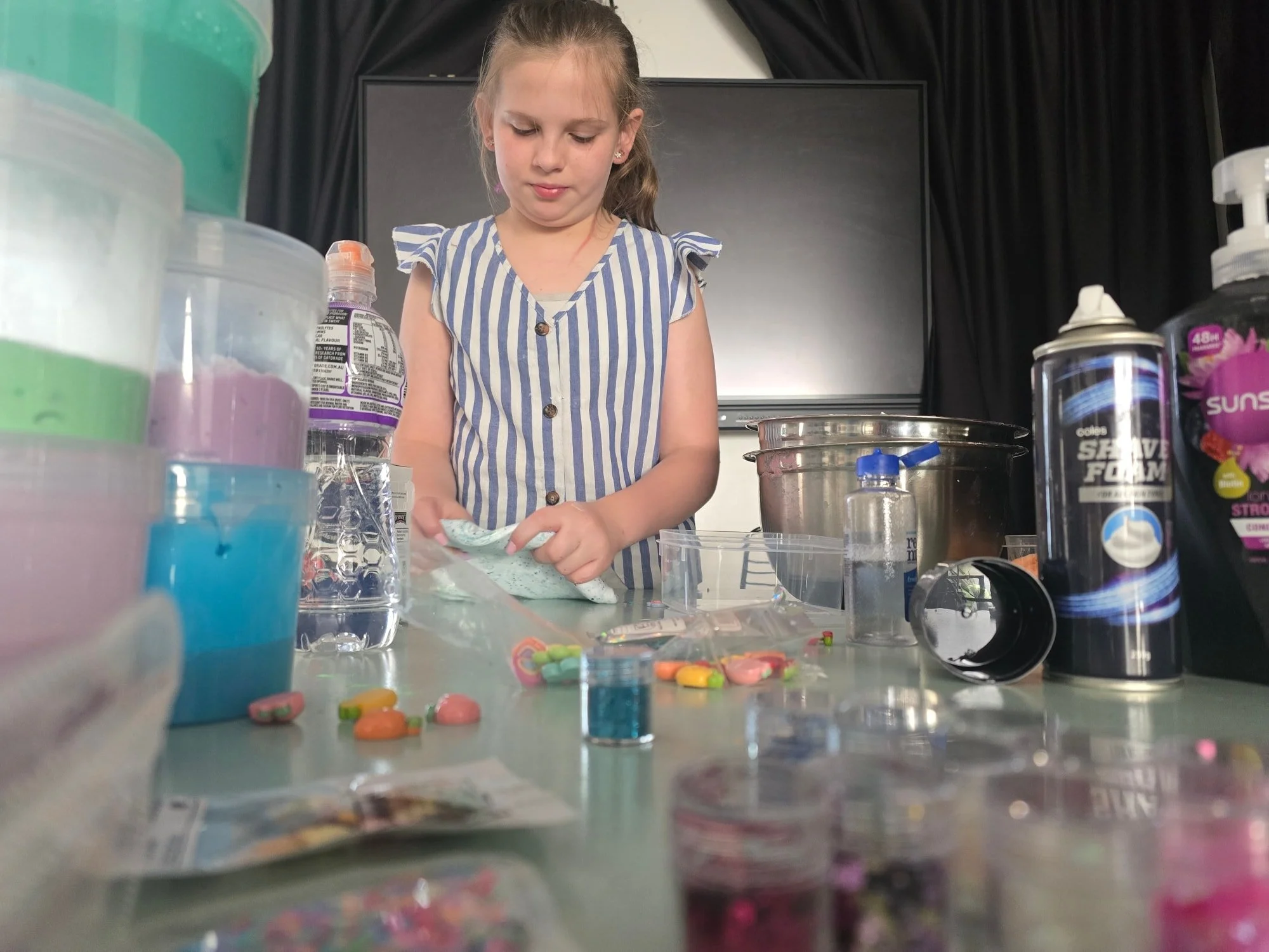 A young girl with blonde hair and a striped shirt is standing at a table, handling colorful candy or pills. The table is cluttered with various containers, bottles, and scattered candy, with a large monitor in the background.