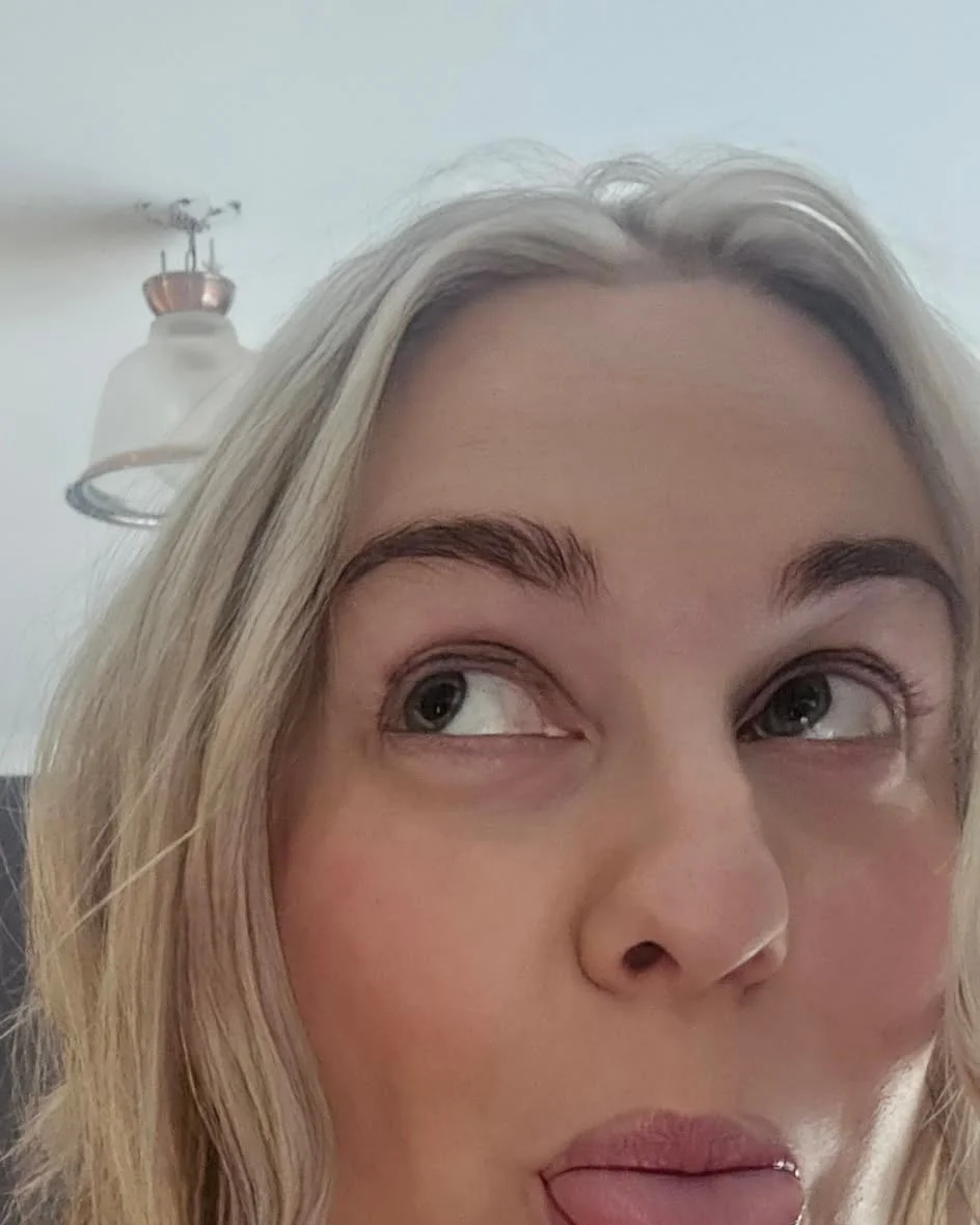 Close-up of a woman with blonde hair sticking out her tongue and looking to the side, with a ceiling fan visible in the background.