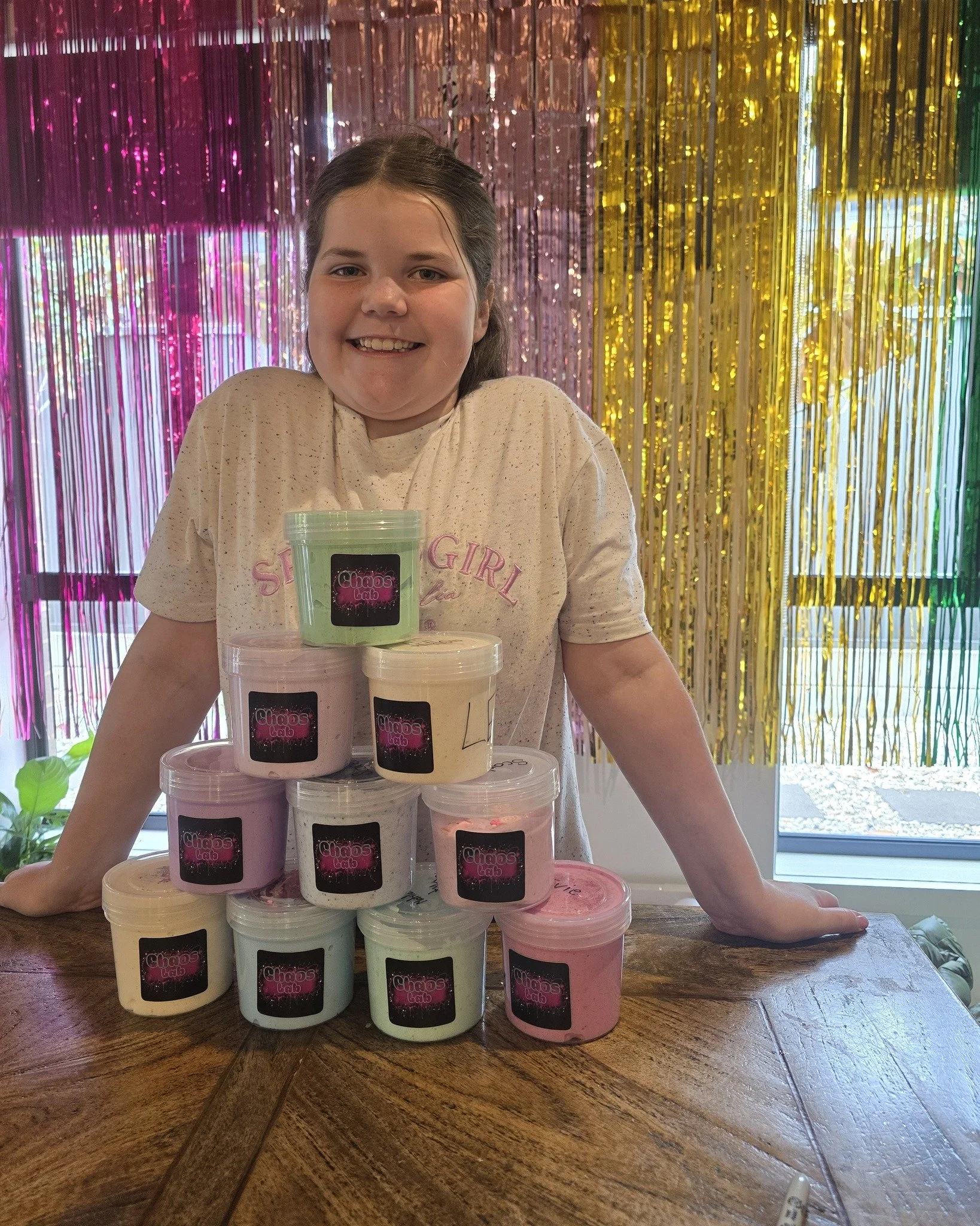 A young girl with brown hair and a light-colored T-shirt, smiling, stands behind a tower of labeled slime containers in various pastel colors on a wooden table, with a colorful metallic curtain in the background.
