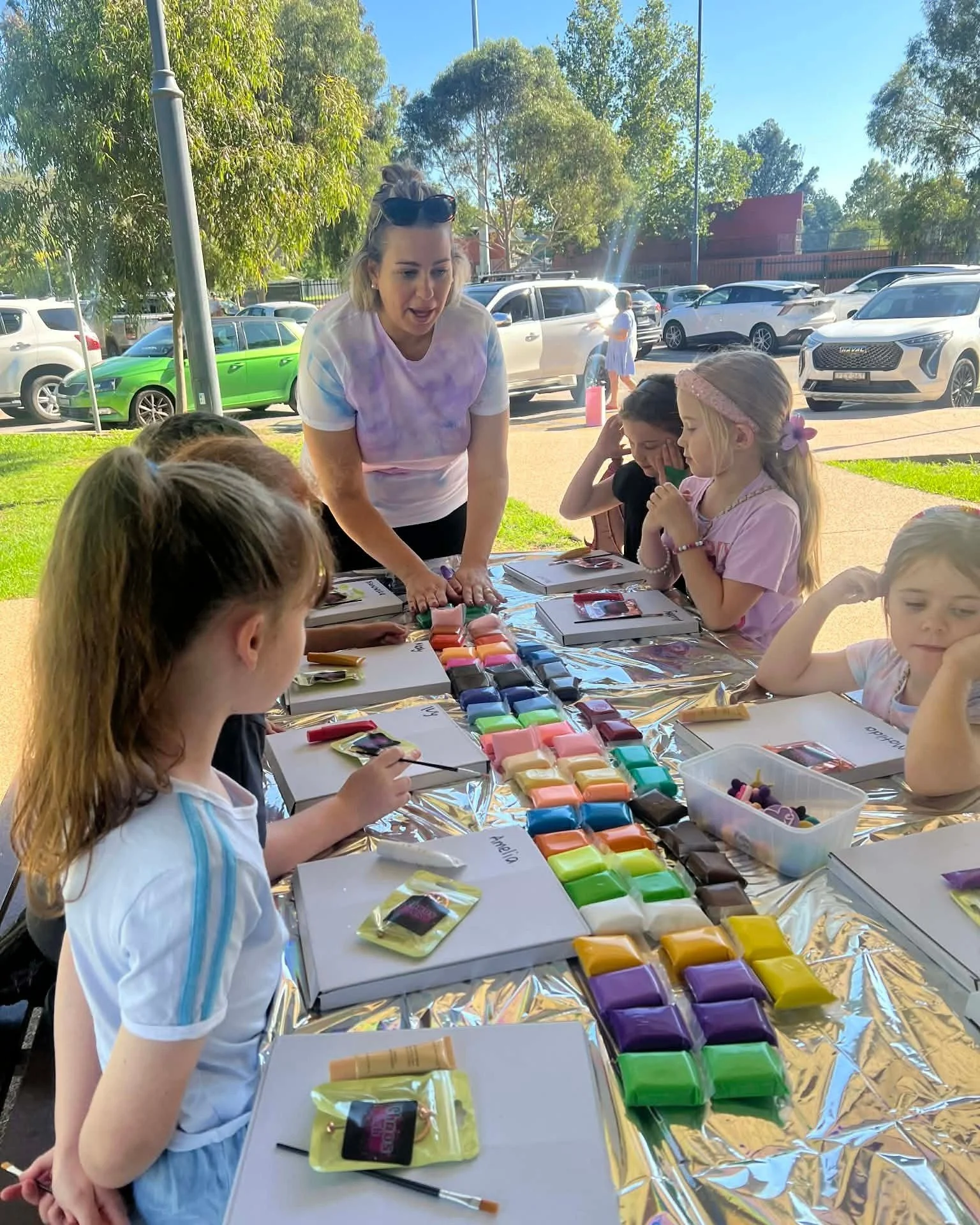 Kids Party.  The table has colorful paint pans and brushes. Behind them, there are parked cars and green trees.