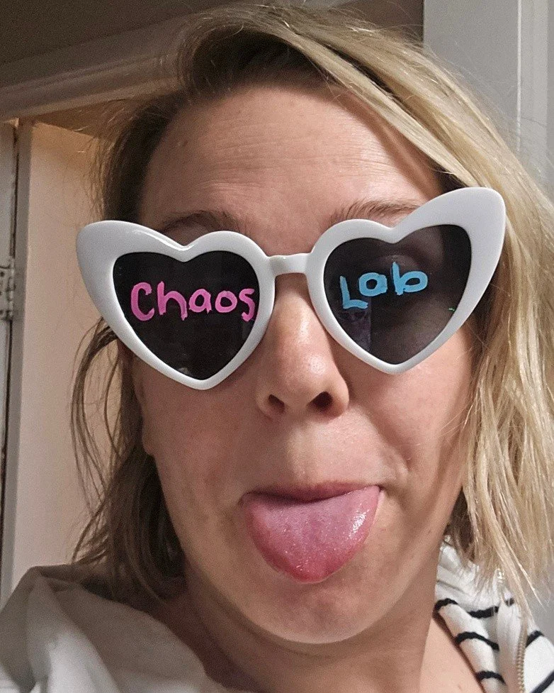 A woman wearing heart-shaped sunglasses with the words 'Chaos' in pink on the left lens and 'Lob' in blue on the right lens, sticking out her tongue.