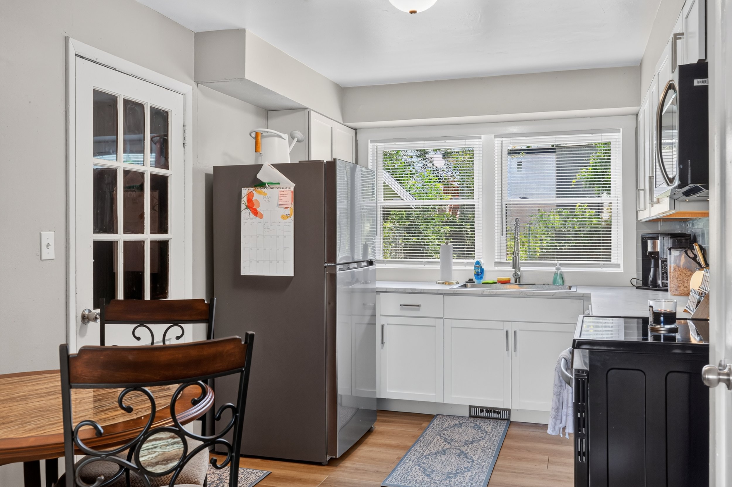 Kitchen with white cabinets, a window with blinds letting in sunlight, a stainless steel refrigerator, a dining table with chairs, and various kitchen items like paper towels, dish soap, and a microwave.
