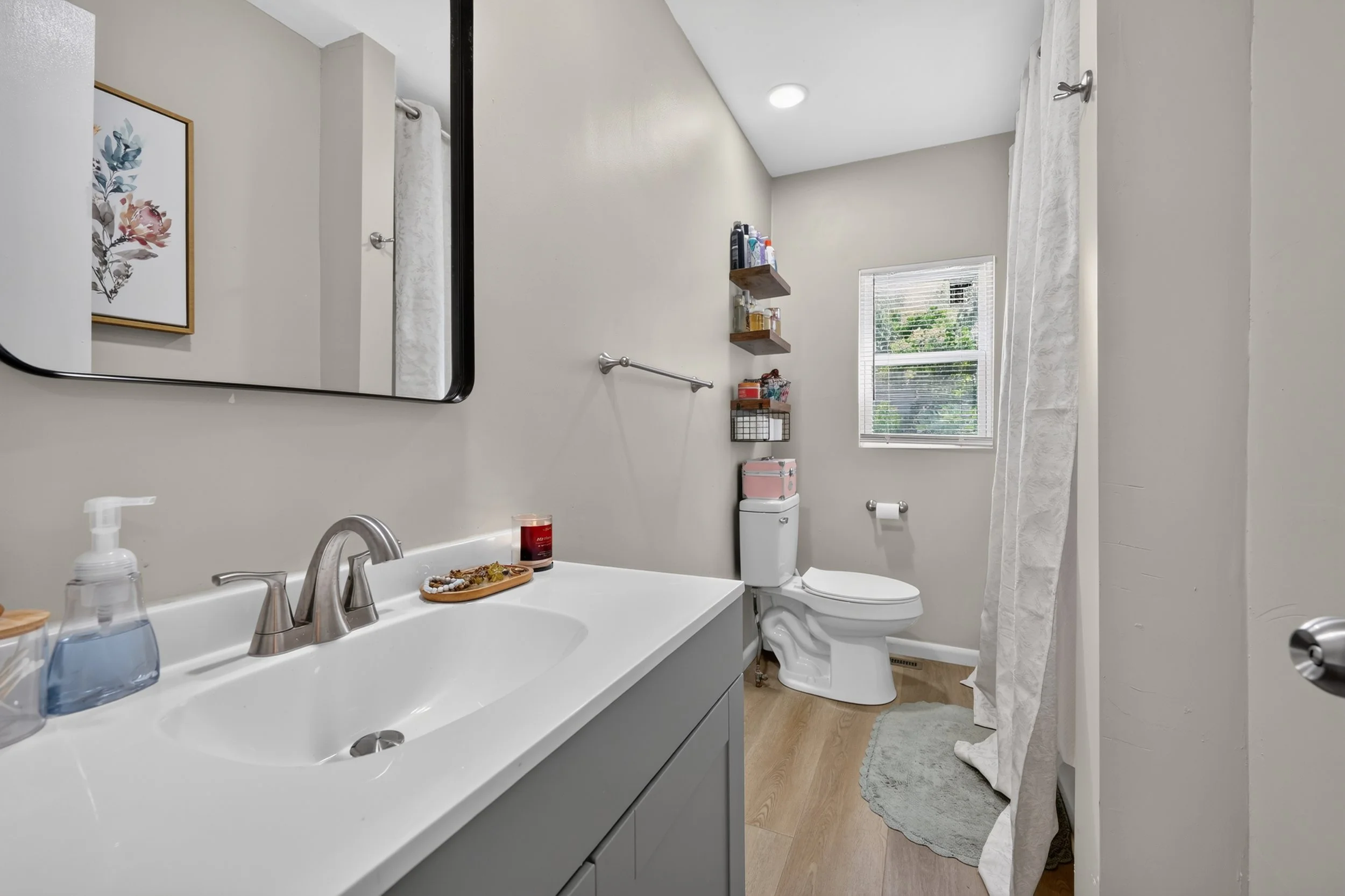 A modern bathroom with a white vanity, a sink with a silver faucet, a soap dispenser, and a plate with decorative items. There is a large mirror, a toilet, and a window with blinds allowing natural light. Shelves with toiletries and a decorative pink box are above the toilet. A shower curtain hangs to the right, and artwork adorns the wall.