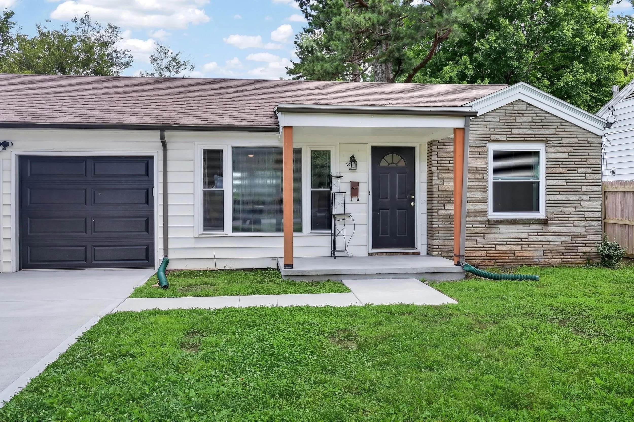 Front exterior of a house with a black garage door, white siding, a small porch with an orange wooden support, and a door with a half-circle window. There's a window with blinds, a mailbox, a small table, and a black lantern on the porch. The grass is green, and trees are in the background.