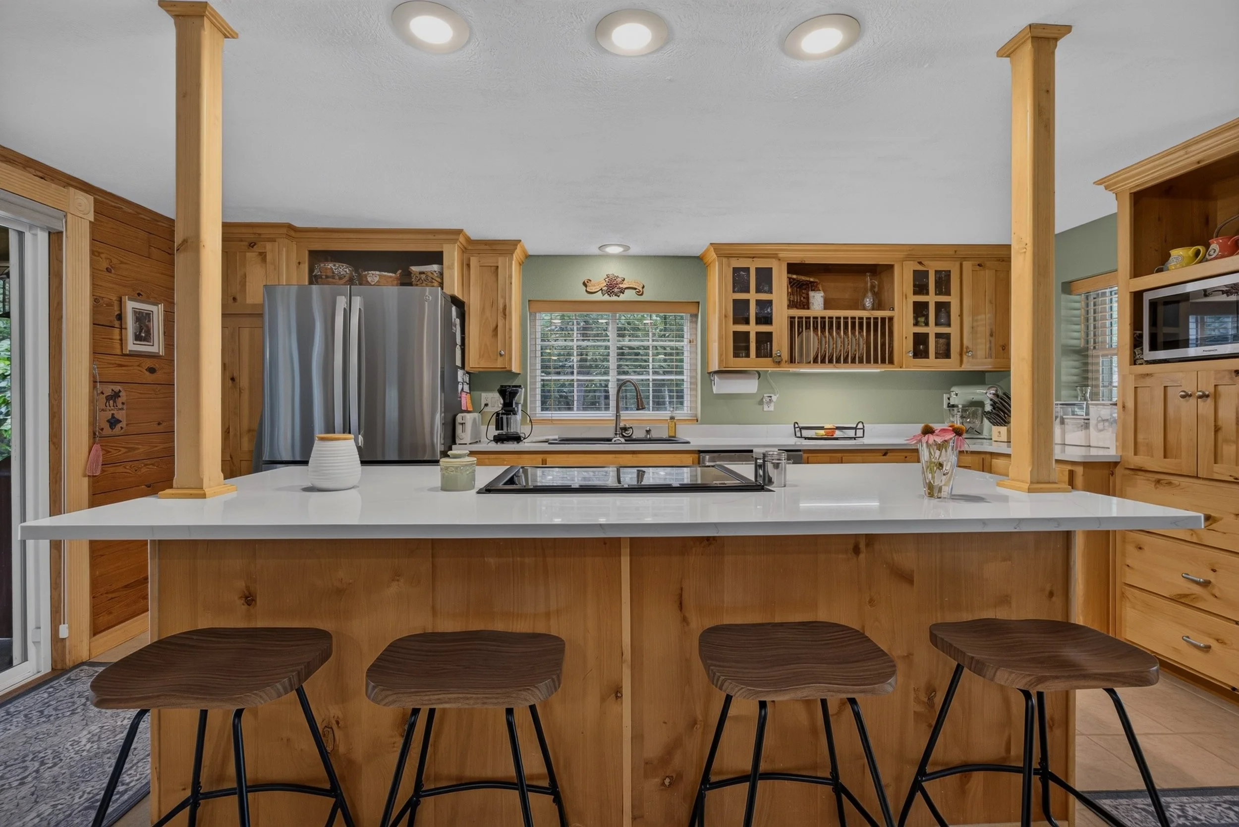 Kitchen with wooden cabinets, stainless steel refrigerator, white countertop, window with blinds, and a kitchen island with three barstools.
