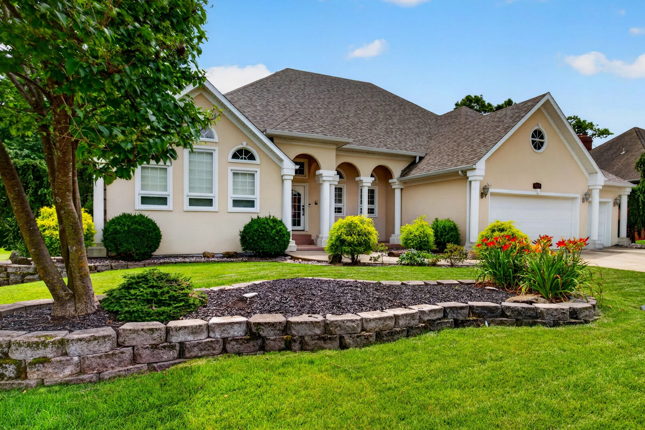 A suburban house with a tan exterior, white trim, and a brown shingle roof. The house has arched windows and columns at the entrance. The front yard features a well-maintained lawn, various shrubs, and a flower bed with red flowers. A tree with green leaves is partially visible on the left side of the image, and the sky is blue with scattered clouds.