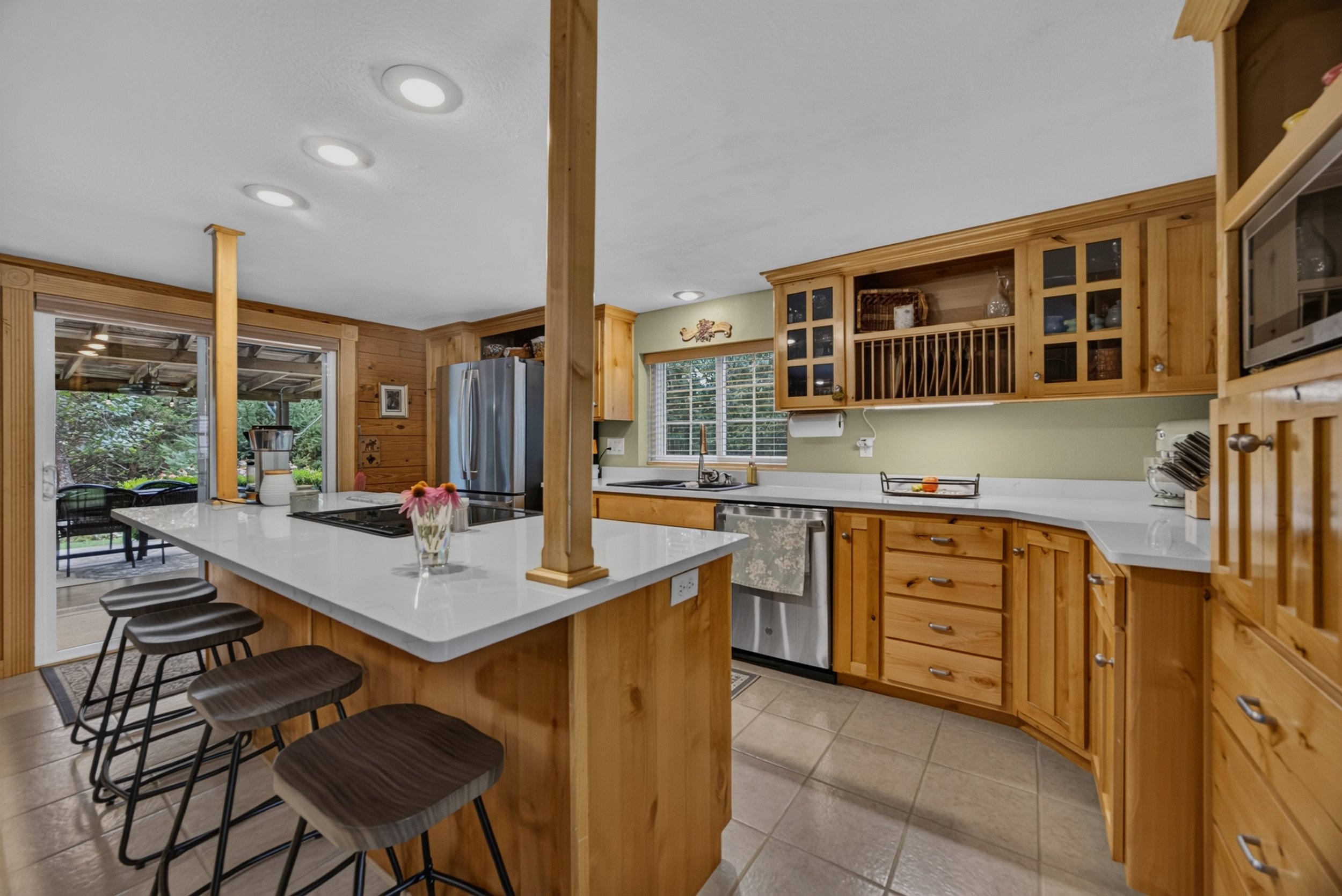 Kitchen with wooden cabinets, a white island with black bar stools, stainless steel appliances, and large windows with views of a patio and green yard.