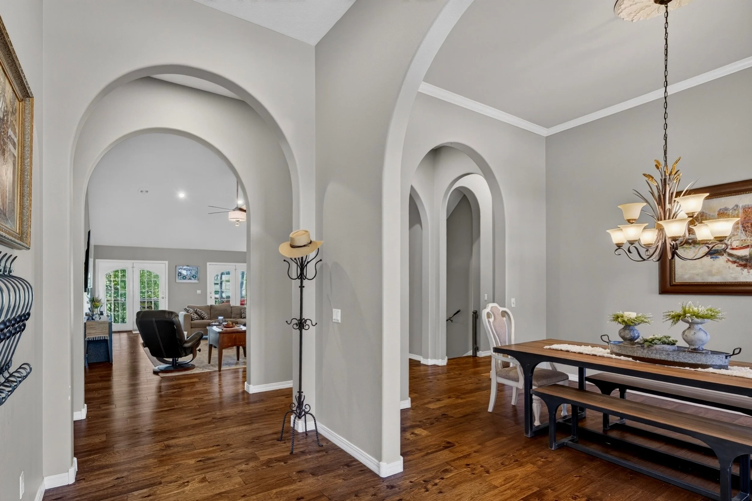 A spacious living room and dining area with high ceilings, white walls, and hardwood floors. Arches divide the spaces, and there is a chandelier hanging over the dining table. Furniture includes a black office chair, a wooden dining table with peeling paint details, and a decorative floor lamp with a hat hanging on it. There are paintings and a door with glass panels, allowing natural light to brighten the room.