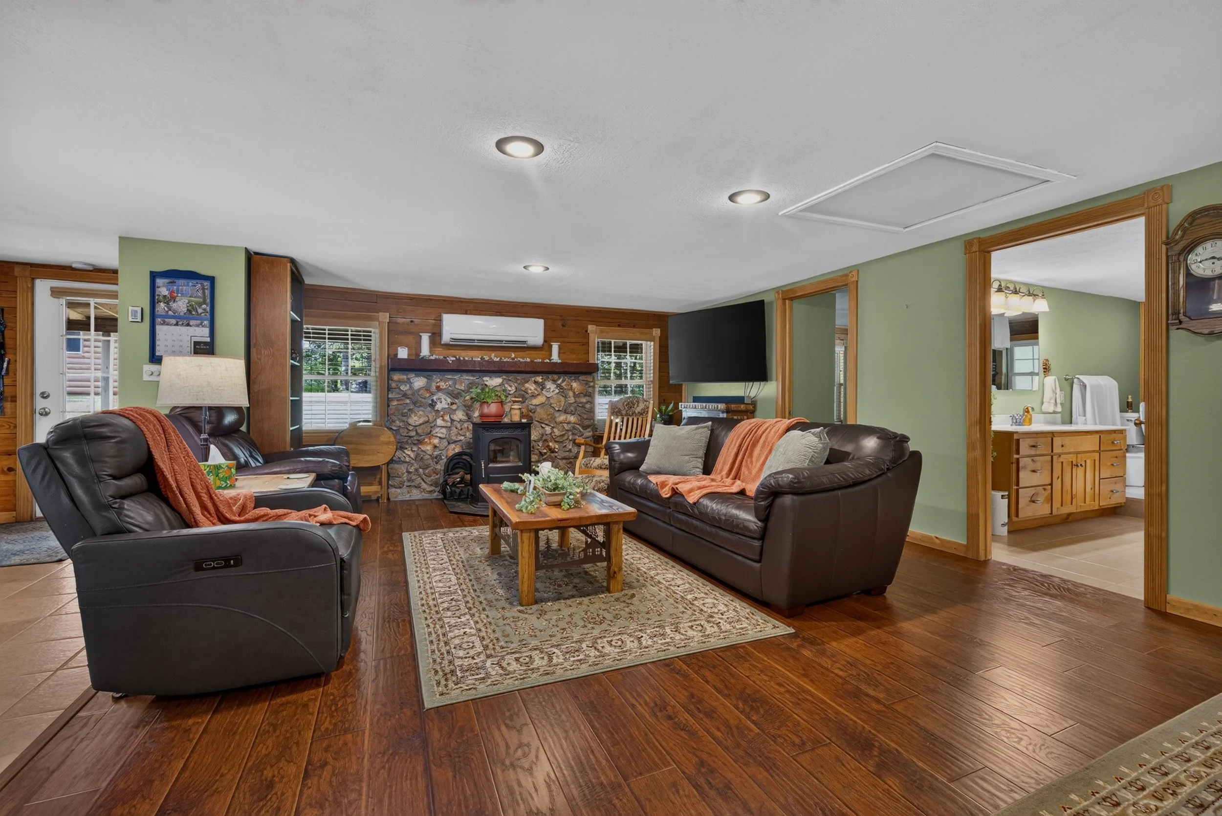 Living room with green walls, hardwood floors, leather sofas with blankets and pillows, a wooden coffee table, a stone fireplace, and a mounted TV, with a view into an adjacent room.
