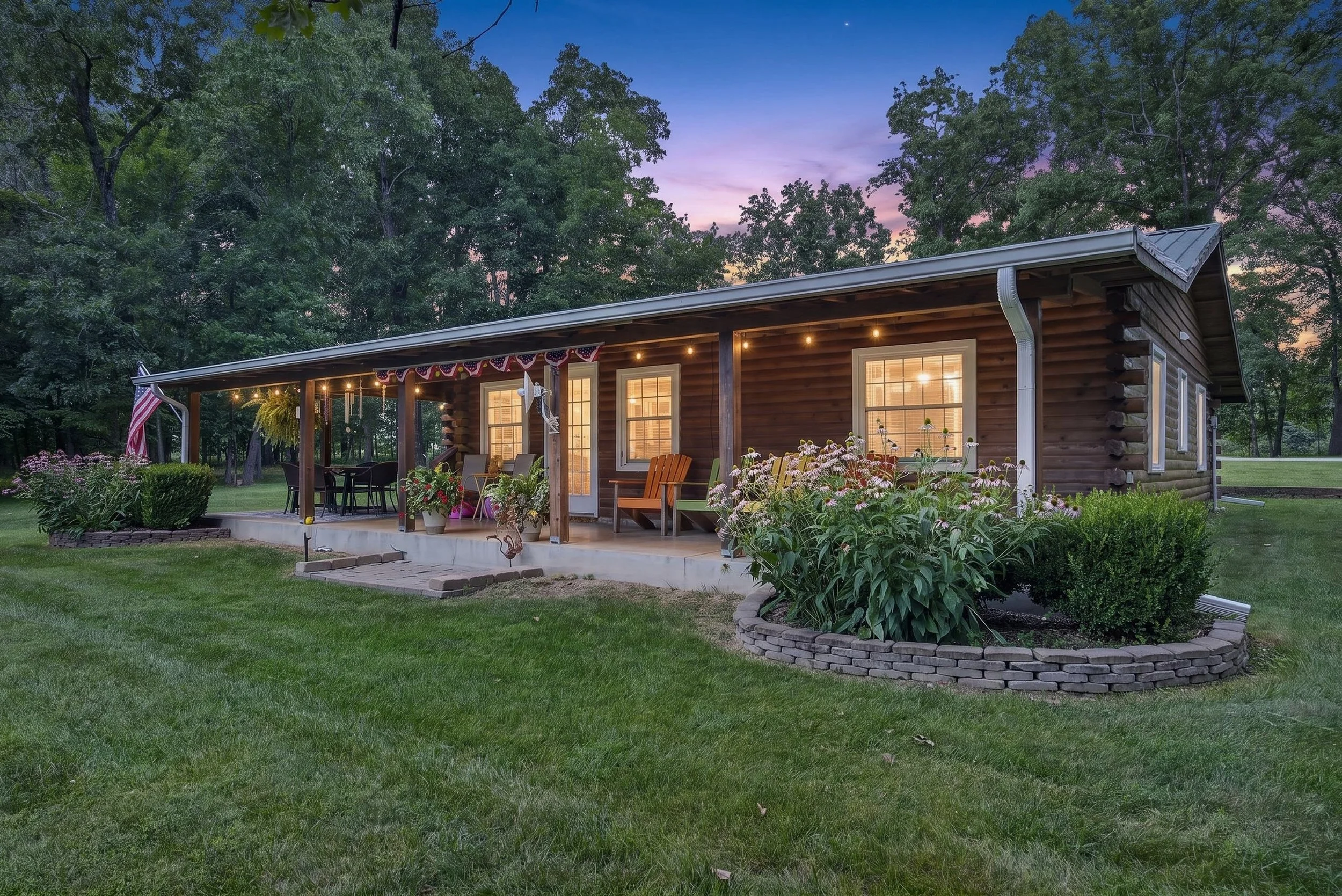 A cozy wooden cabin with lit windows and a porch decorated with string lights and American flags. There are outdoor chairs and potted plants on the porch, surrounded by a well-maintained garden and lush green lawn, with tall trees and a colorful sunset sky in the background.