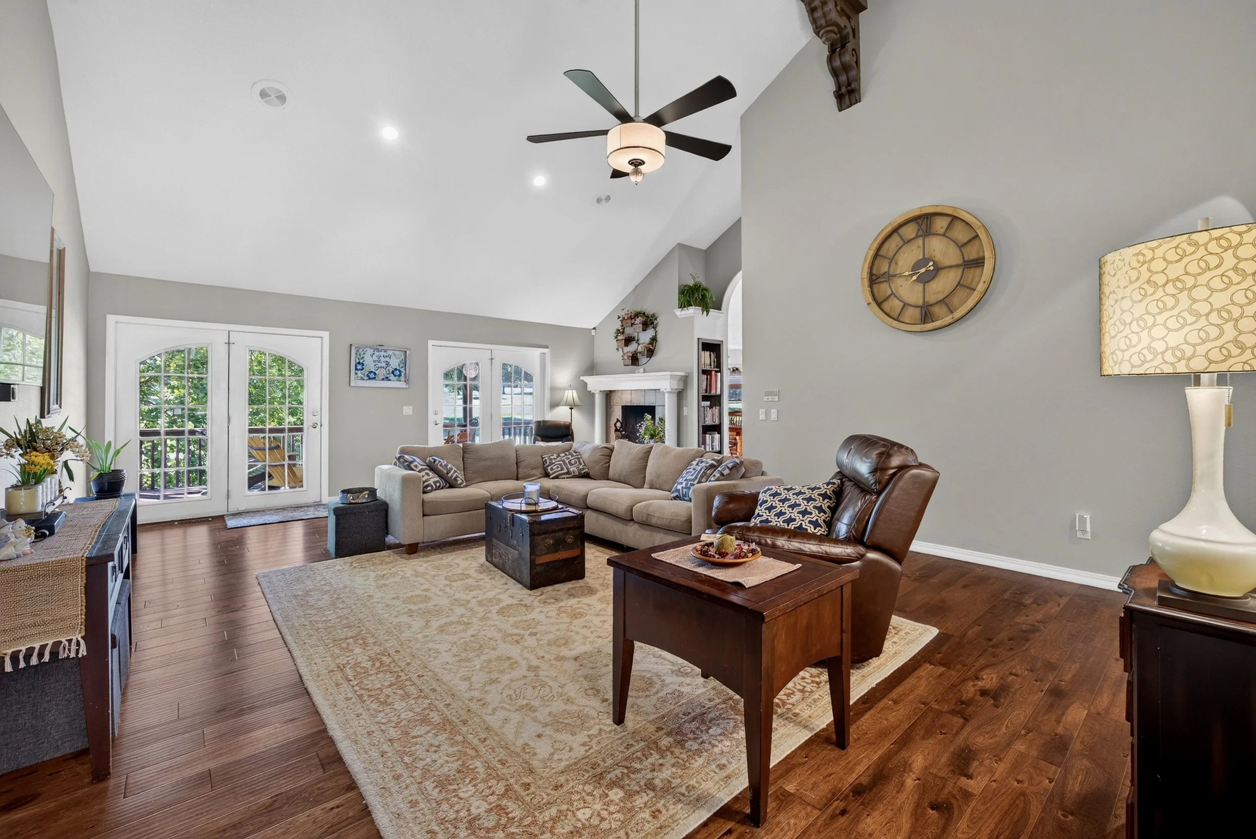 Living room with a beige sectional sofa, a wooden coffee table, and a leather armchair. A fireplace with a mantle and a bookshelf are visible. Double glass doors lead outside, and a ceiling fan with lights is overhead. The room has gray walls and hardwood floors.