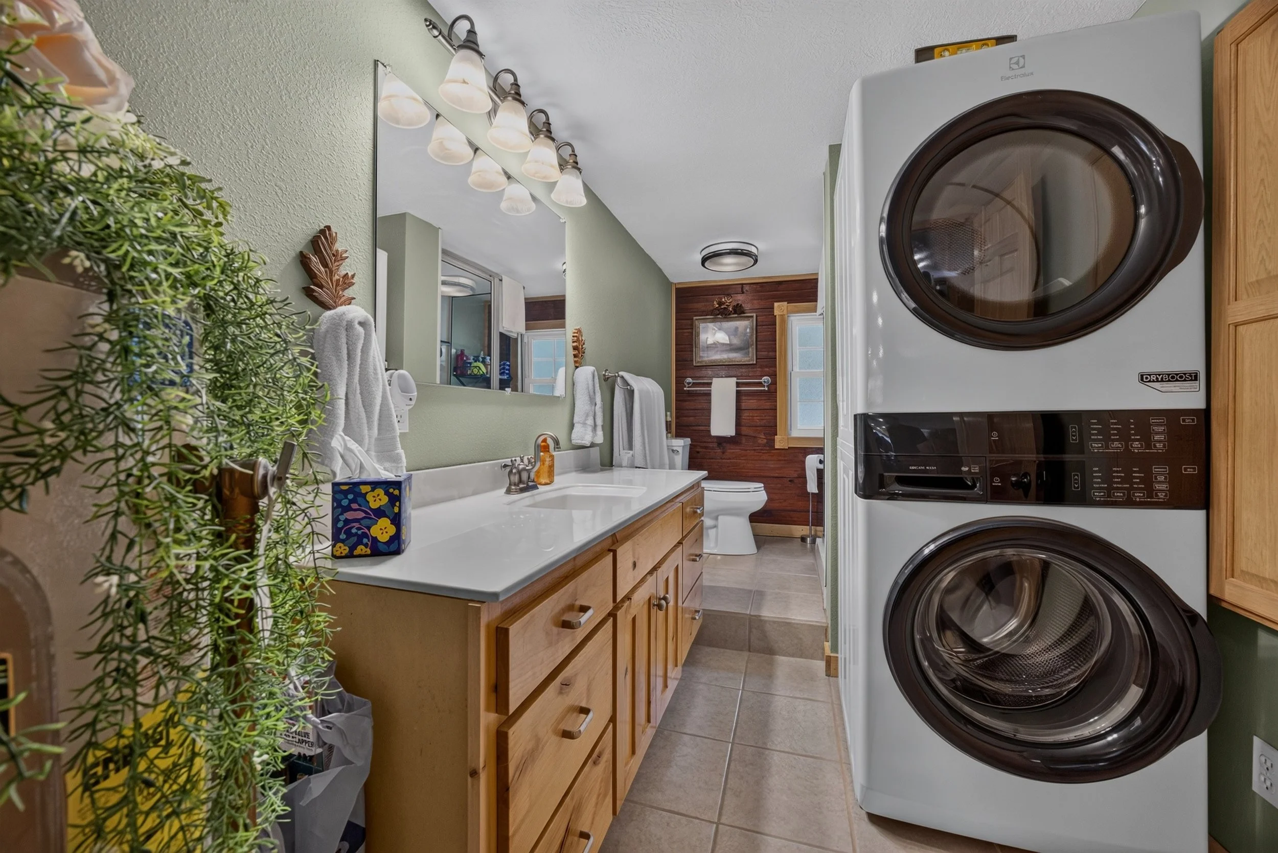 A laundry room with a washer and dryer stacked on the right, a wooden vanity with a sink and a mirror above it, various towels, plants, and a box of tissues on the countertop, and a wood-paneled wall with a framed picture in the background.