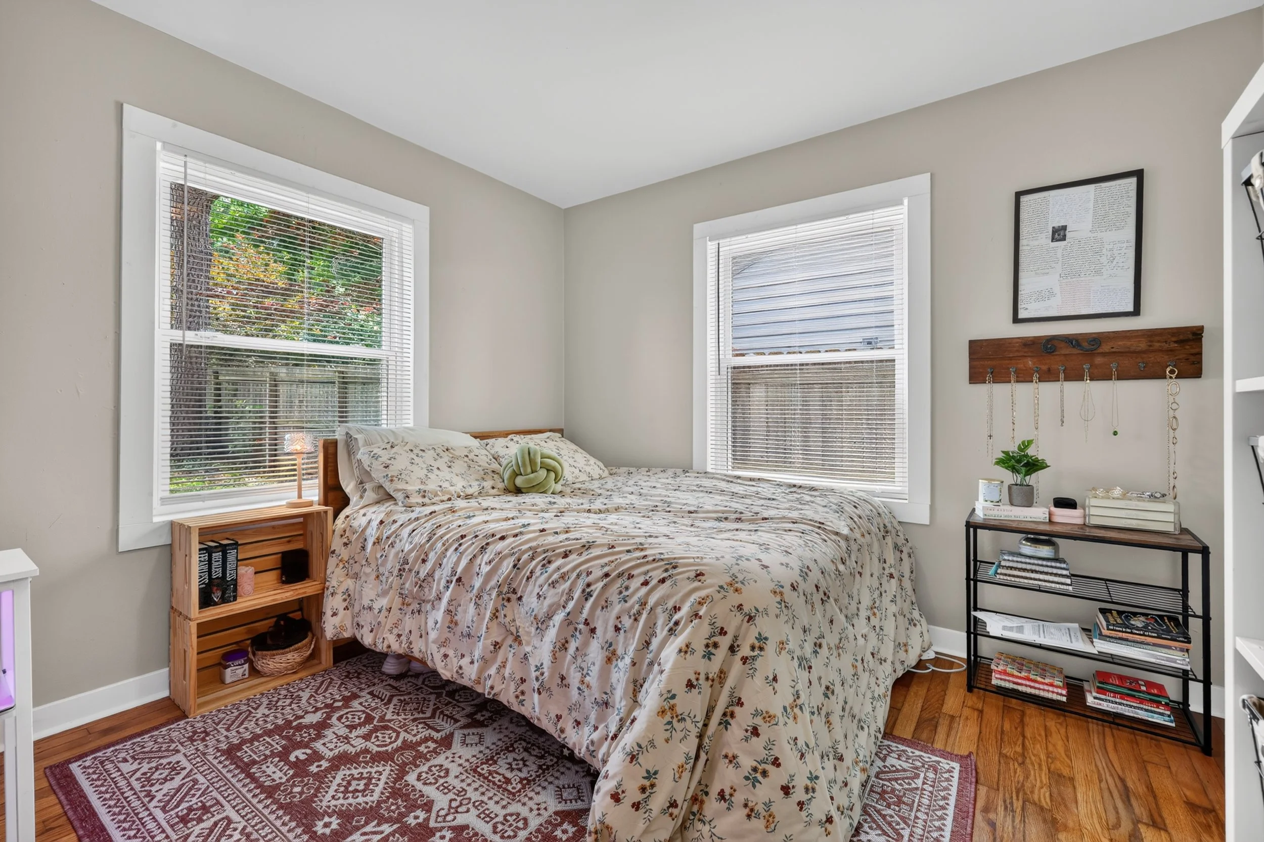 A cozy bedroom with two large windows, a bed with floral bedding, a small wooden nightstand, and a black metal shelf filled with books and decor. There is a framed print and a wooden jewelry organizer on the wall, and a plant on the shelf.