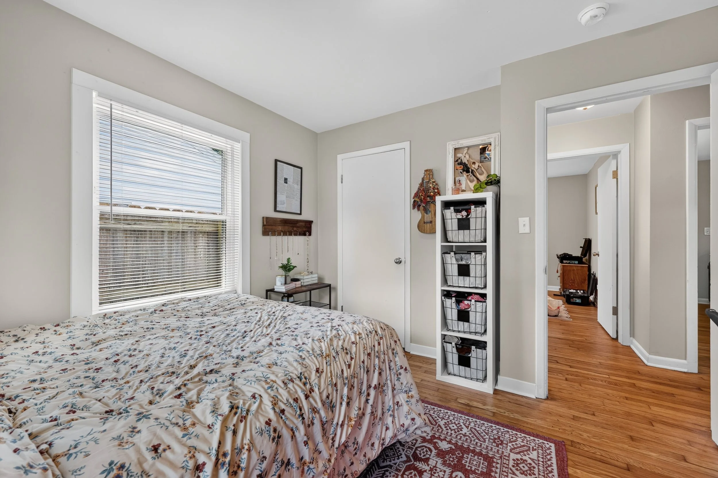 A cozy bedroom with a window, framed with white trim, letting in natural light. There is a bed with floral bedding, a small table with books and a plant, and a white storage unit with baskets. The room has light-colored walls, hardwood floors, and a patterned area rug. Adjacent to the bedroom is a hallway with open doorways.
