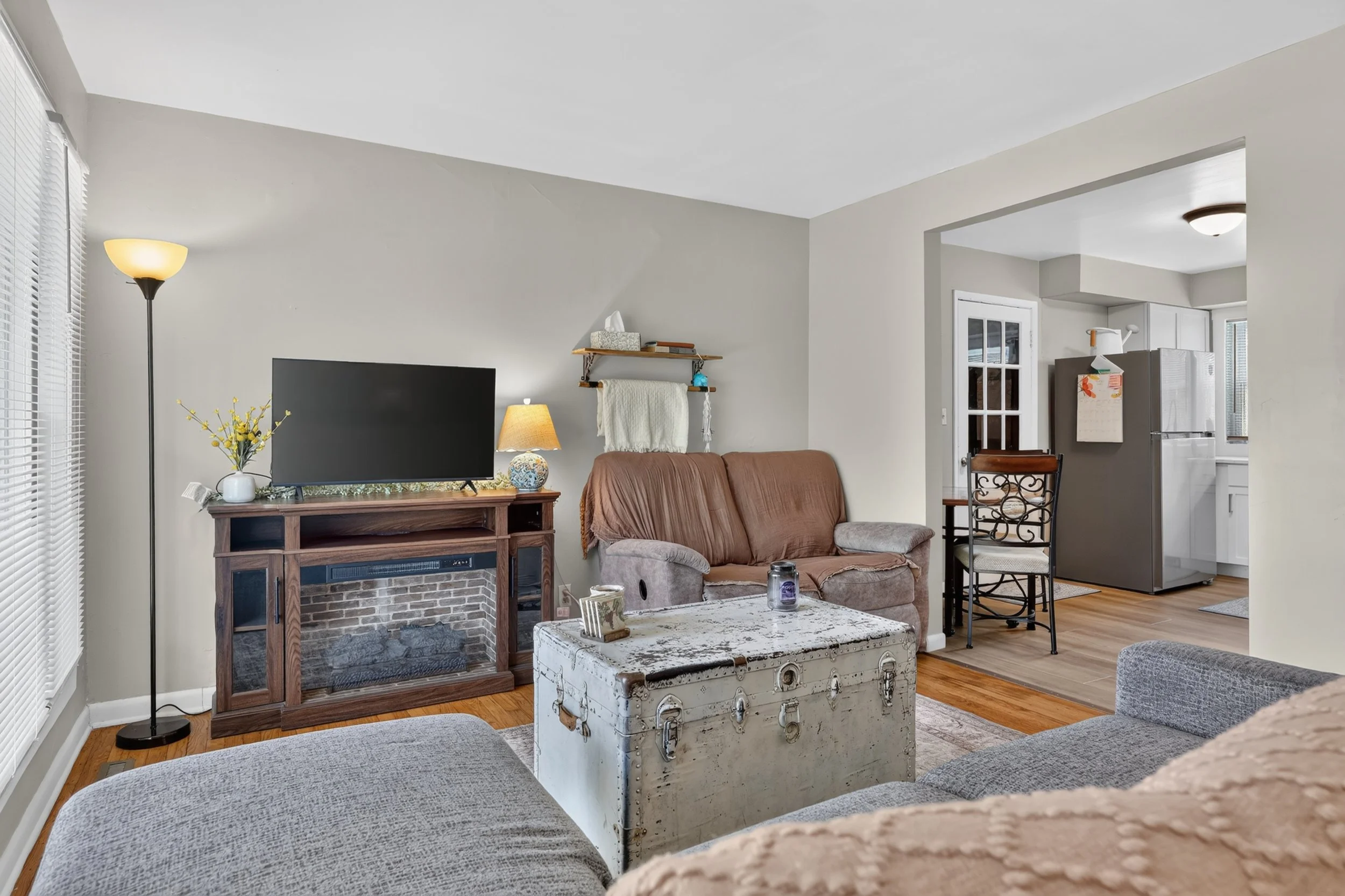 Living room with beige walls, wooden floor, and a large window with blinds. Contains a brown sofa, a vintage white trunk, a wood fireplace with a TV on top, a tall floor lamp, a small side table with a floral lamp, and decorative items on a wall shelf.