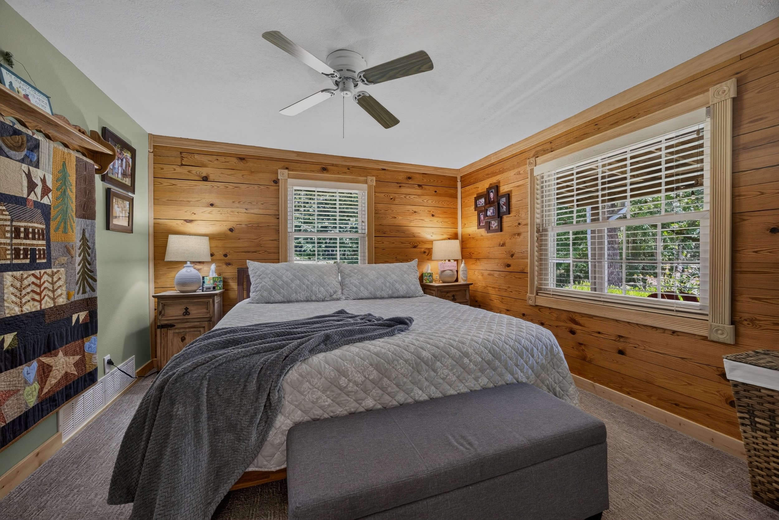 A cozy bedroom with wooden panel walls, a bed with white quilt and gray blanket, two bedside lamps, a quilted ottoman at the foot of the bed, and windows with white blinds showing green trees outside.
