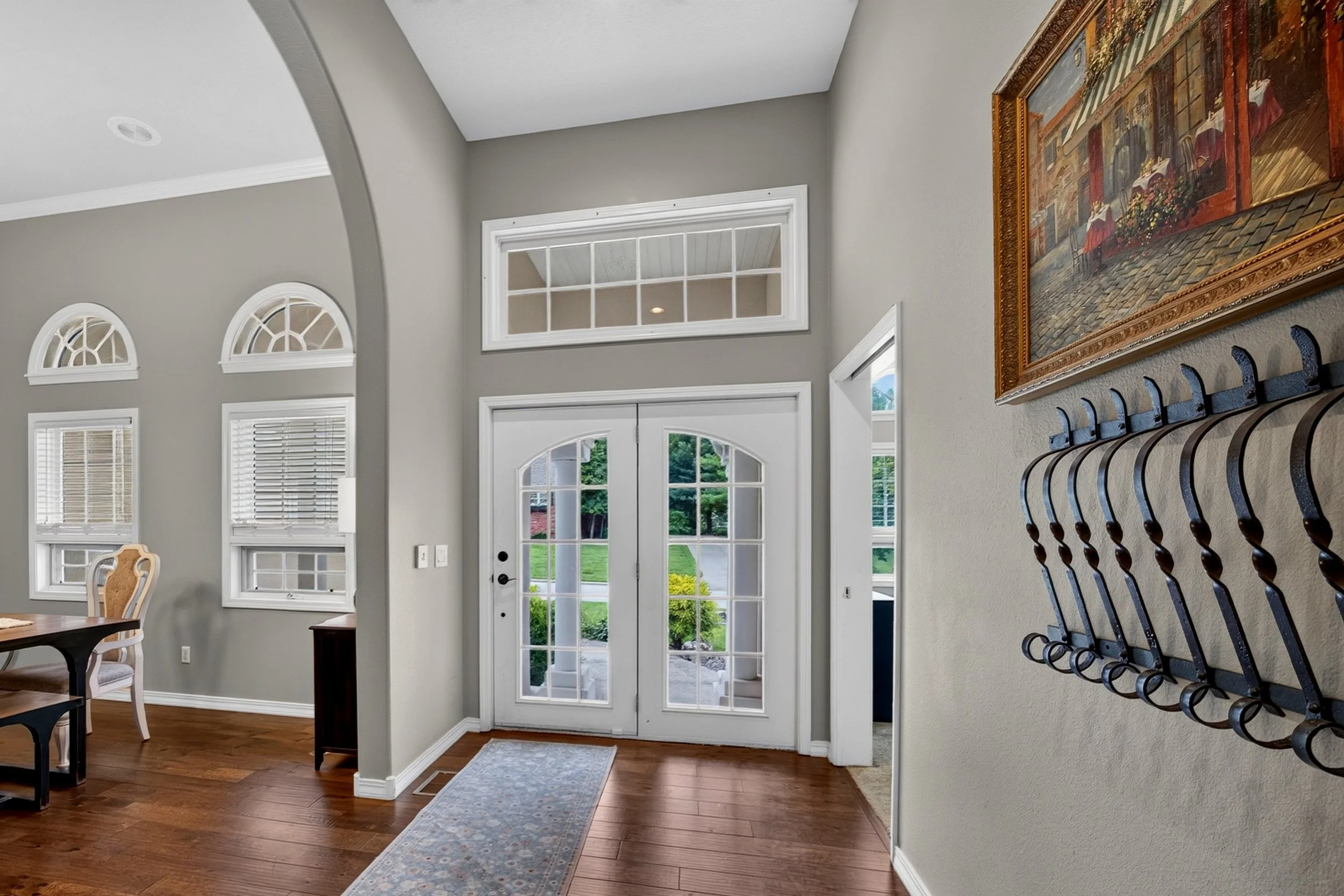 Inside view of a residential entryway with glass double doors, a hallway with windows and a wall with a framed painting, a coat rack, and a dining area with a chair and table.