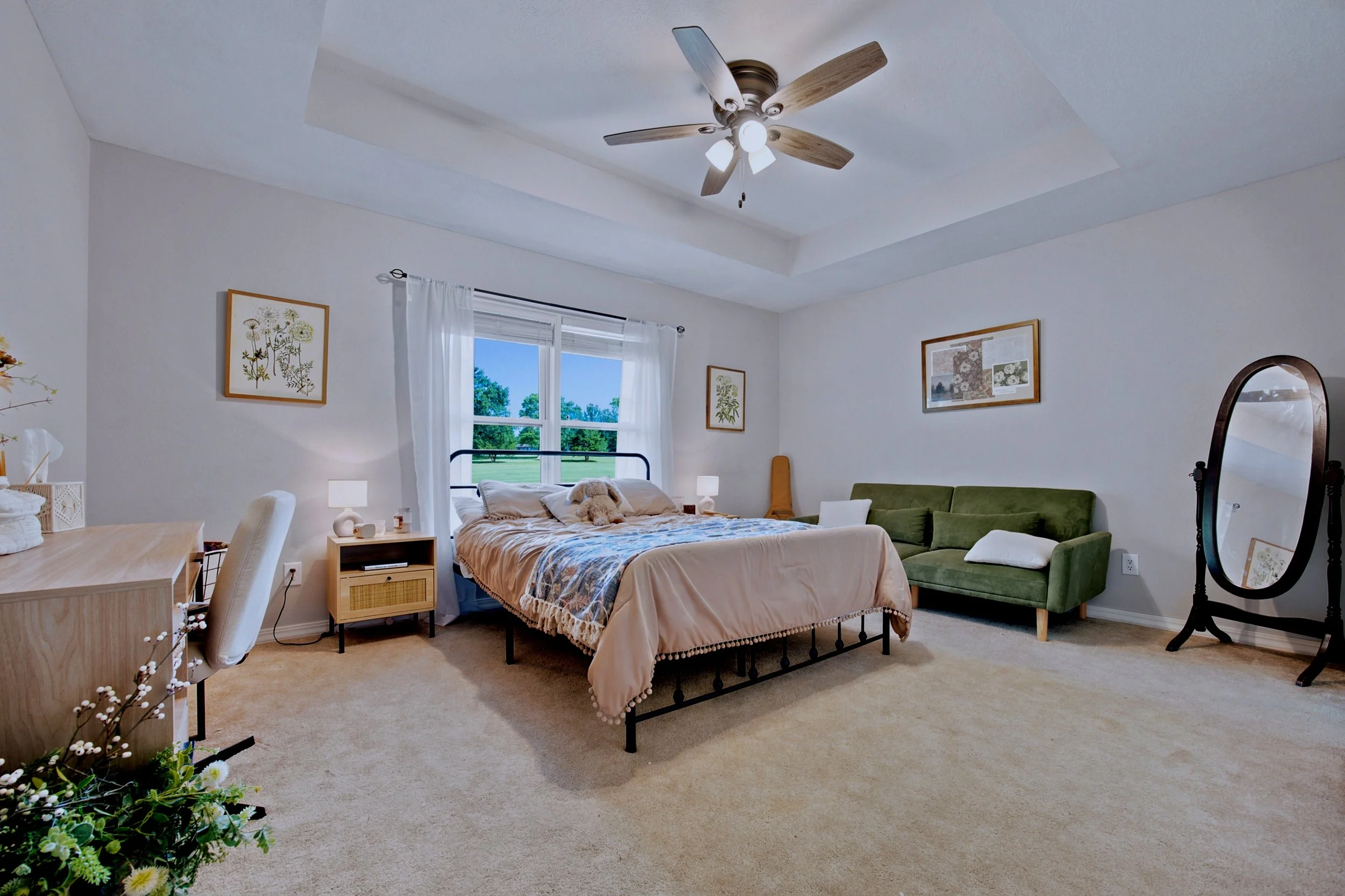 A bedroom with a bed, green sofa, and a large window showing trees outside. The walls are decorated with framed artwork, and there are lamps beside the bed and a mirror on a stand.