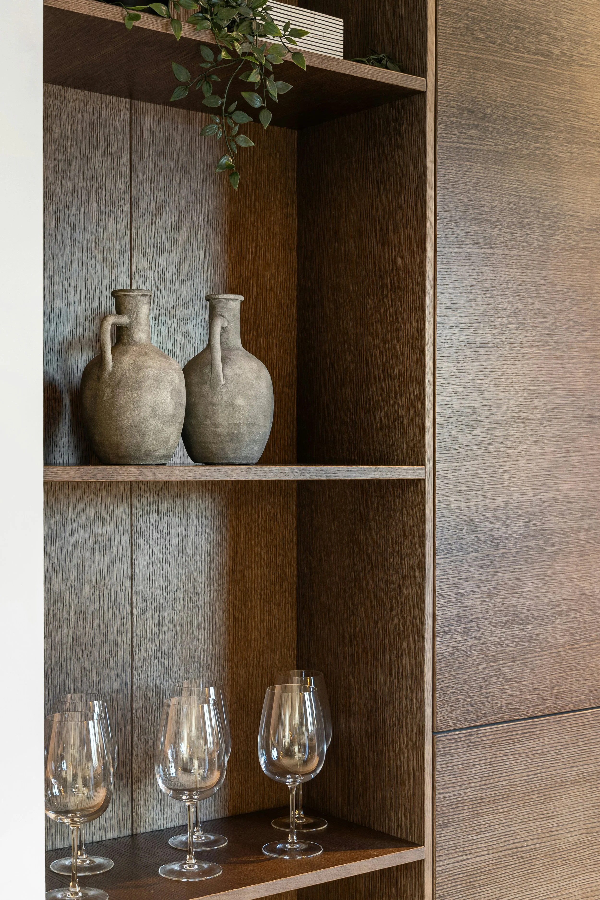 Wooden shelves with two beige ceramic vases with handles on a lower shelf and four empty wine glasses on a counter below.