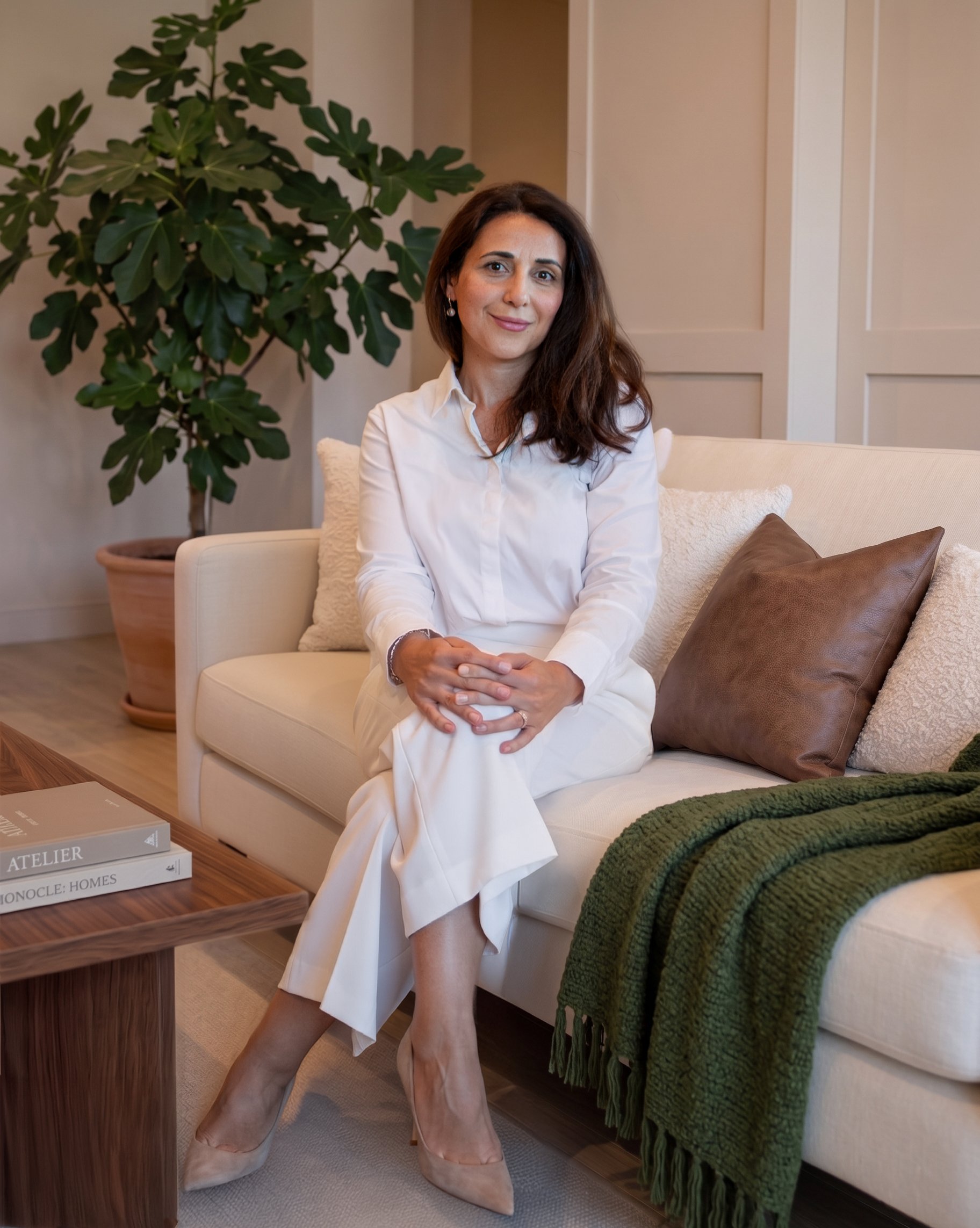 A woman with dark brown hair wearing a white dress and beige heels sitting on a beige sofa in a living room, with a large green houseplant behind her, beige paneled wall, a dark brown pillow, and a green throw blanket on the sofa.