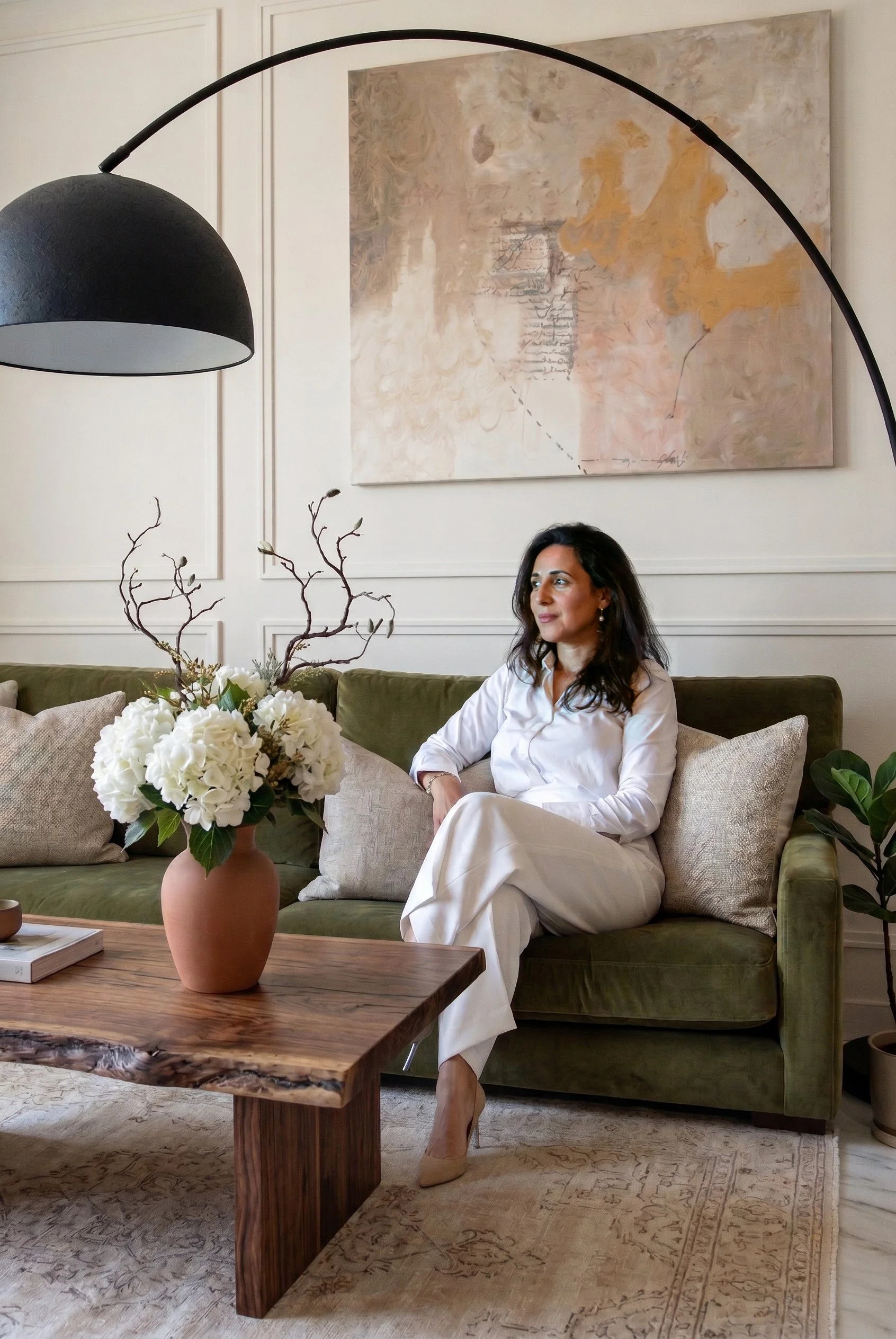 A woman sitting on a green sofa in a living room with beige walls and abstract artwork. There is a wooden coffee table with a terracotta vase holding white flowers and branches in the foreground.