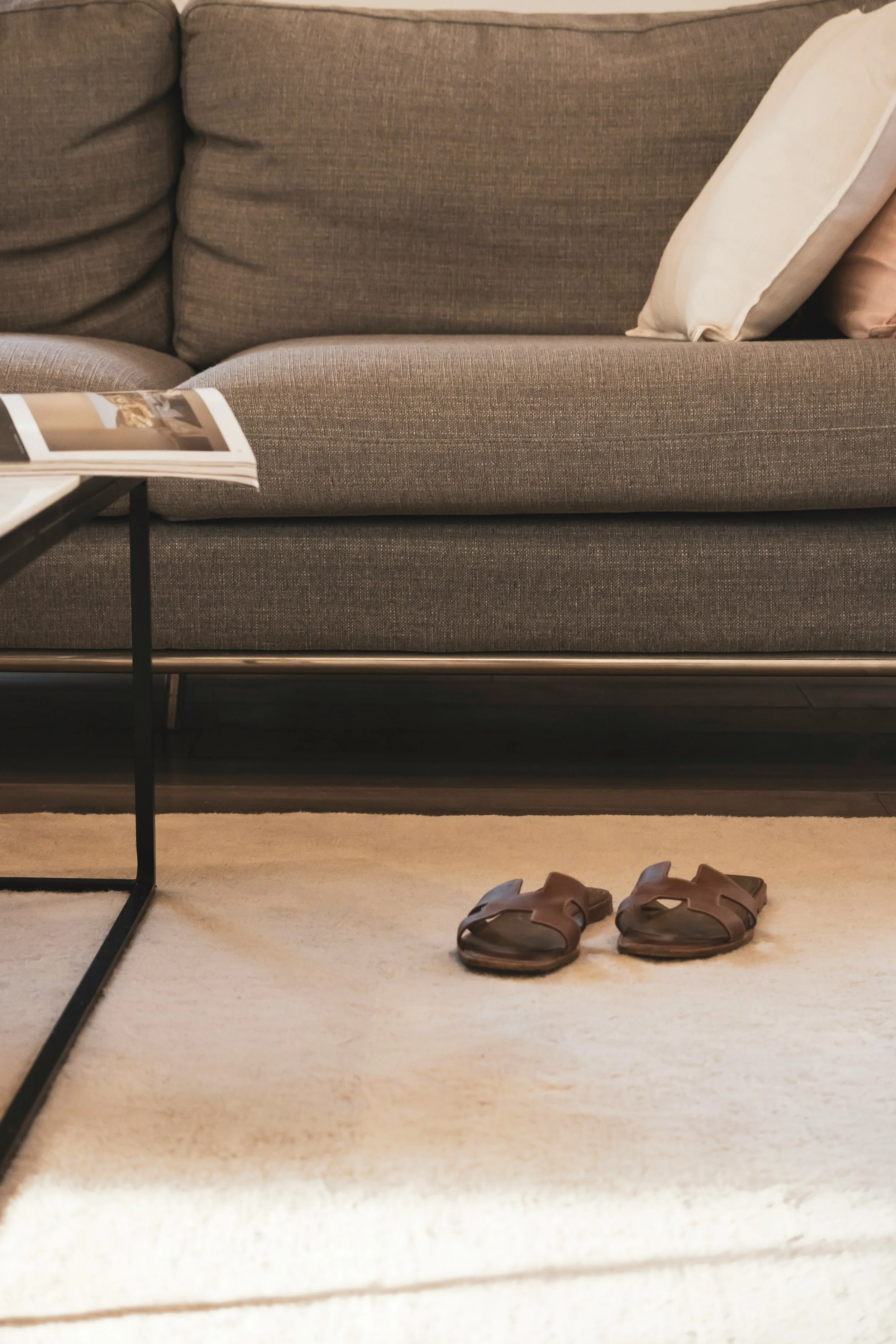 A pair of brown sandals on a cream-colored carpet in front of a gray sofa with beige and pink pillows. A black metal side table with a magazine on top is partially visible on the left.