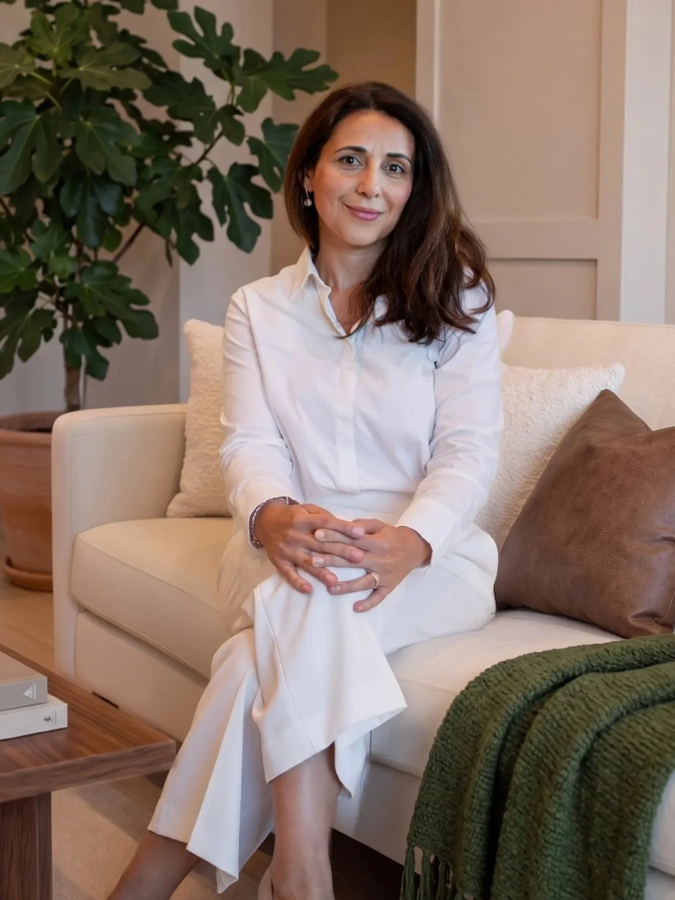 A woman with dark brown hair sitting on a beige sofa with her hands on her lap, wearing a white shirt and cream-colored pants, smiling in a living room with a large green houseplant and decorative pillows.