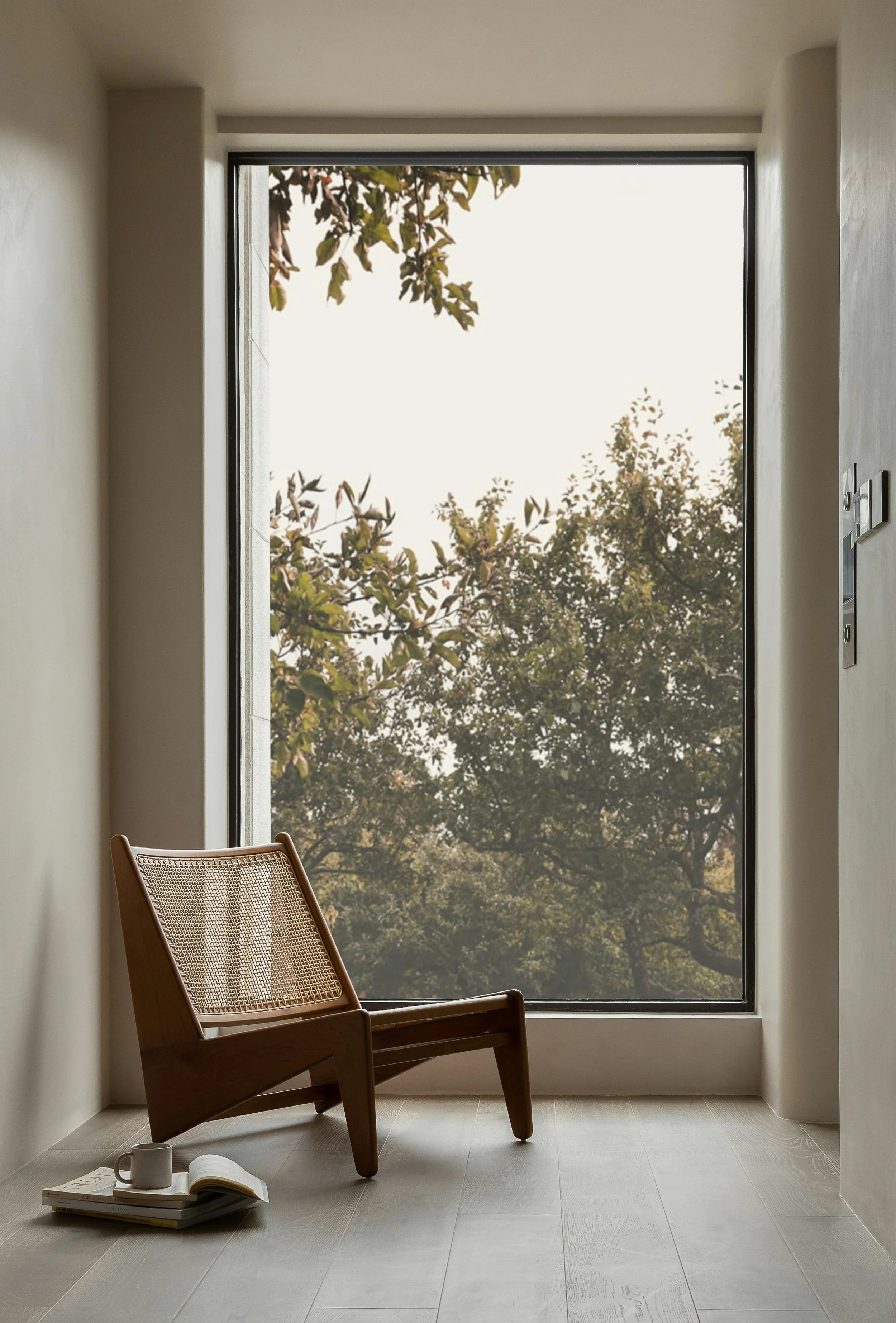 Wood chair, stack of books, open notebook, mug, and teabag on a white floor in front of a large window showing trees outside through sheer curtains.