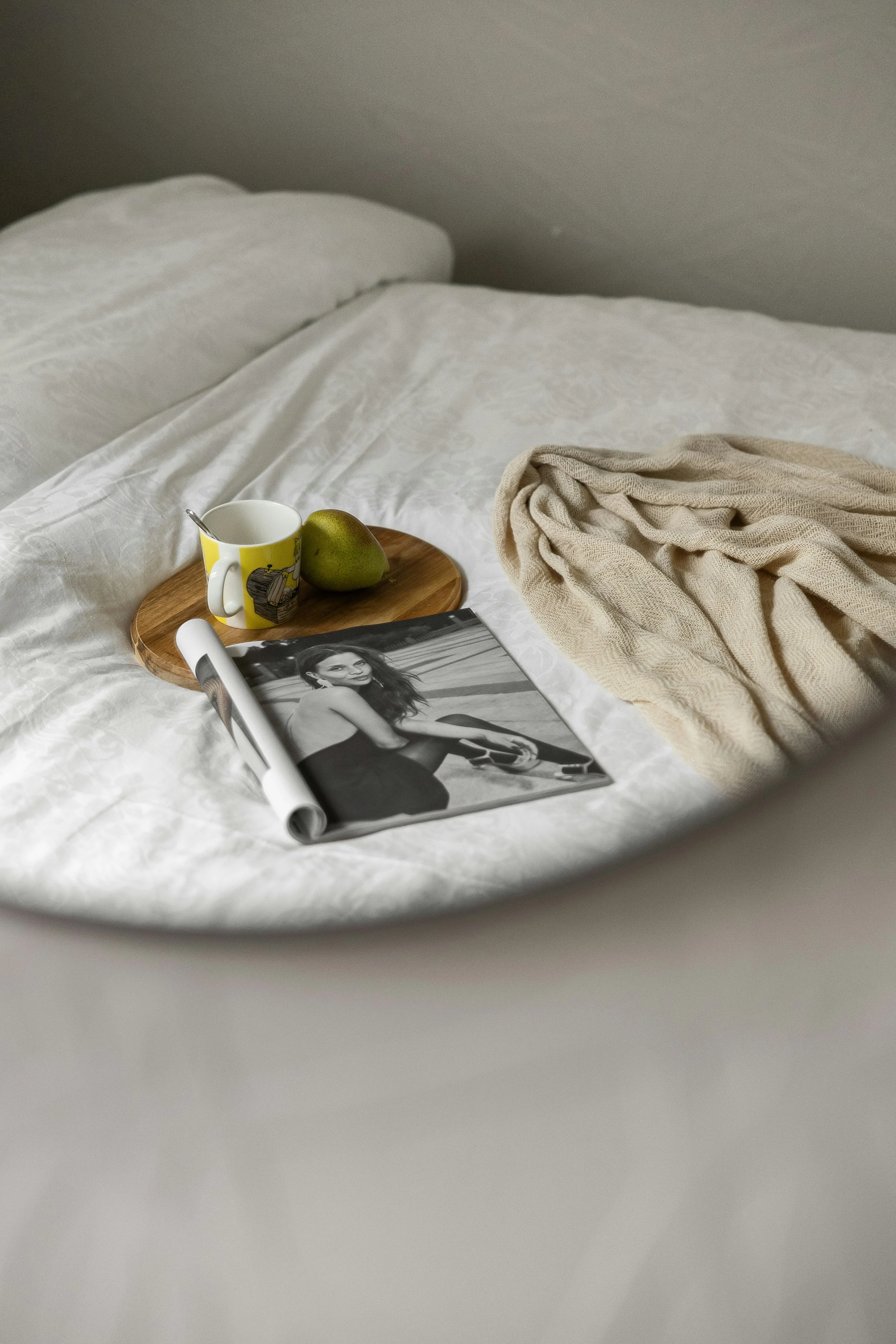 A white bed with a beige blanket, a yellow mug with a spoon, a green pear, a rolled-up magazine, and a black and white magazine featuring a woman in a black dress.