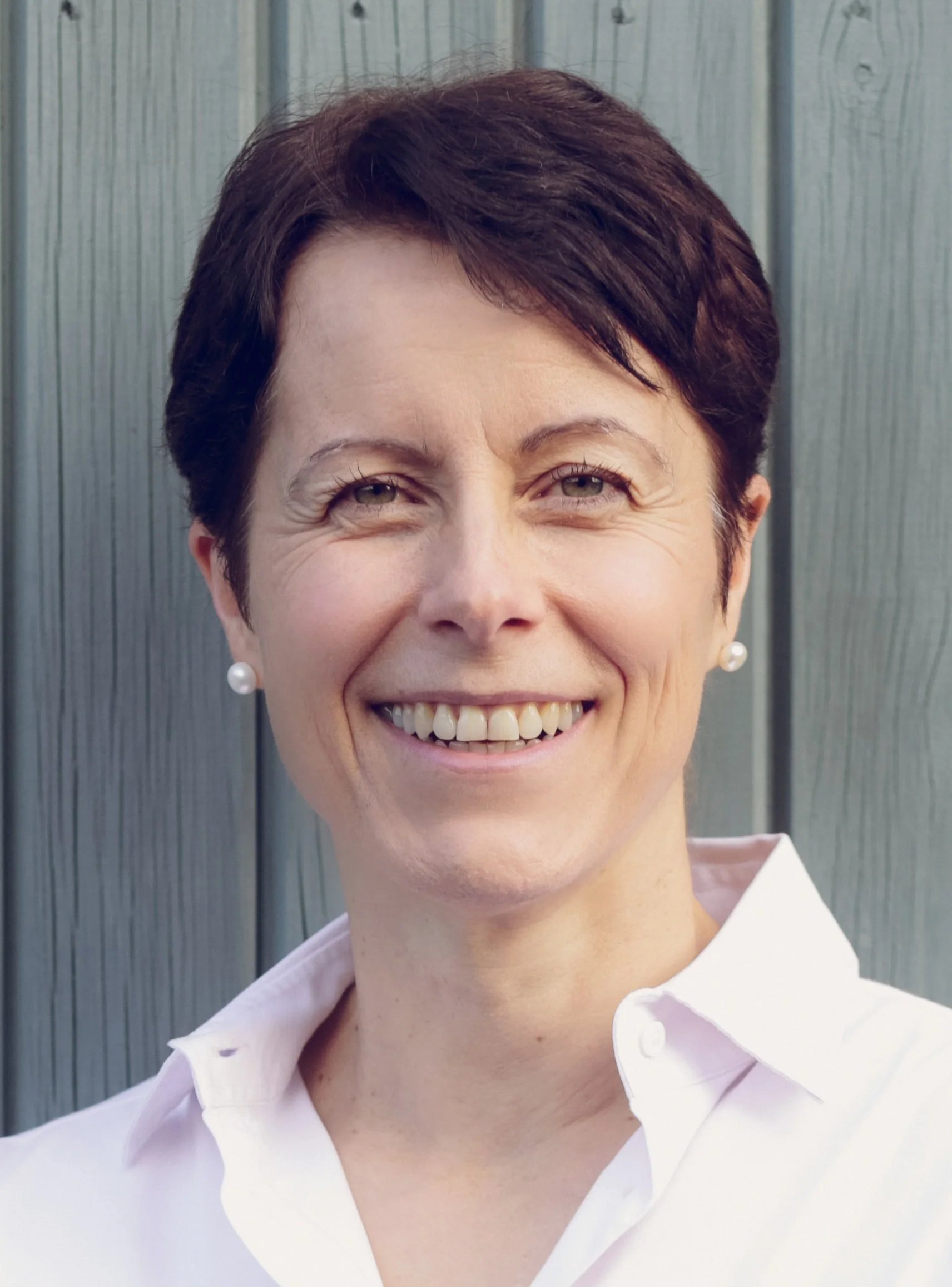 Close-up of a smiling woman with short dark hair, wearing pearl earrings and a white collared shirt, standing outdoors against a gray wooden background.