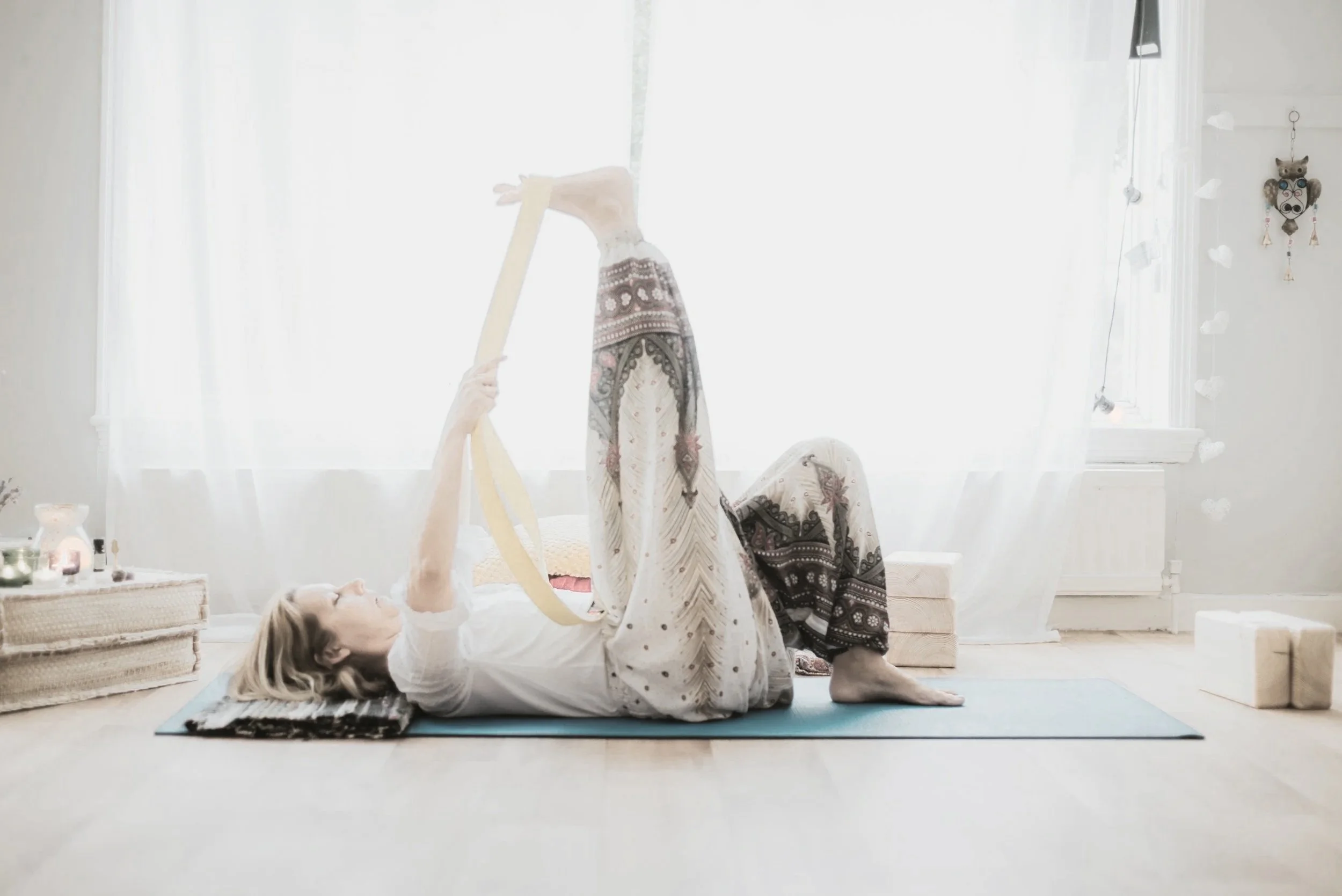 An elderly woman practicing yoga indoors, lying on her back with her legs elevated and she is holding her feet with her hands, on a yoga mat in a bright room with white curtains and a decorated wall.