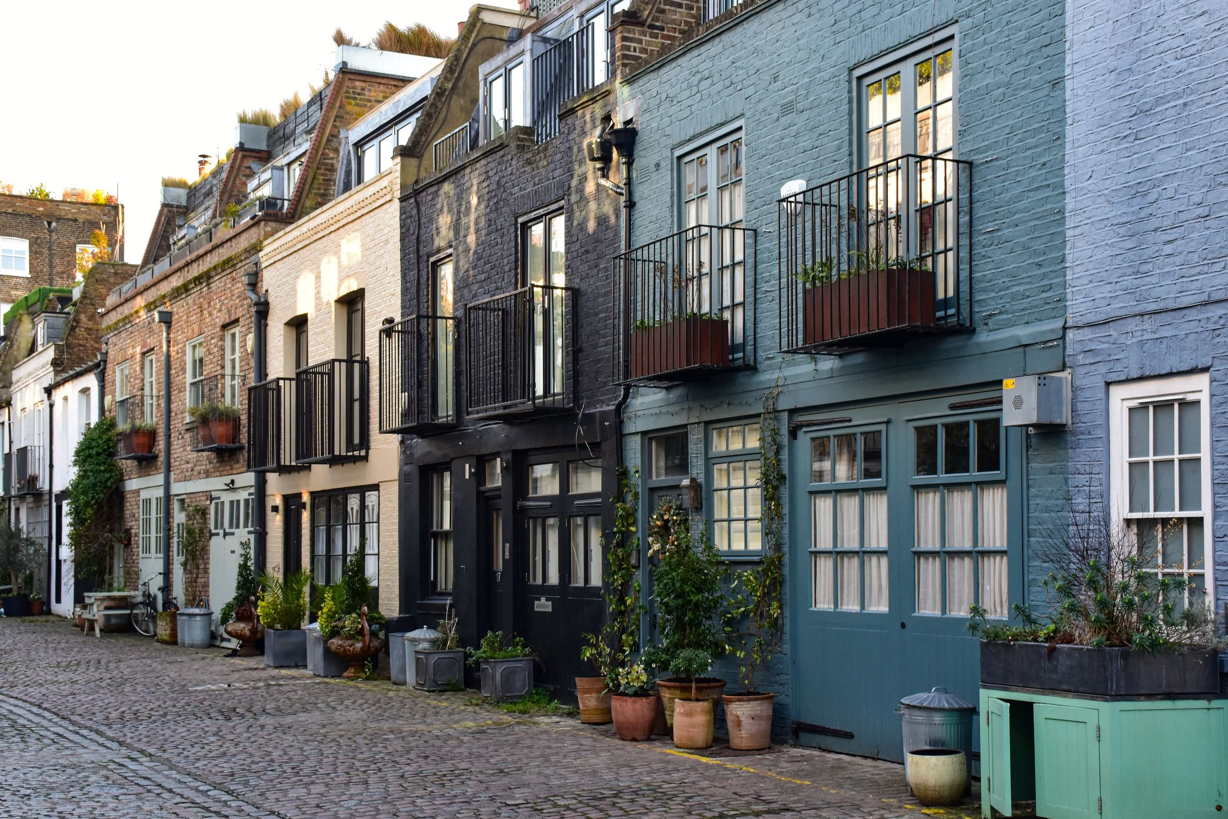 Row of colorful residential buildings with small balconies, potted plants, and a cobblestone street in front.