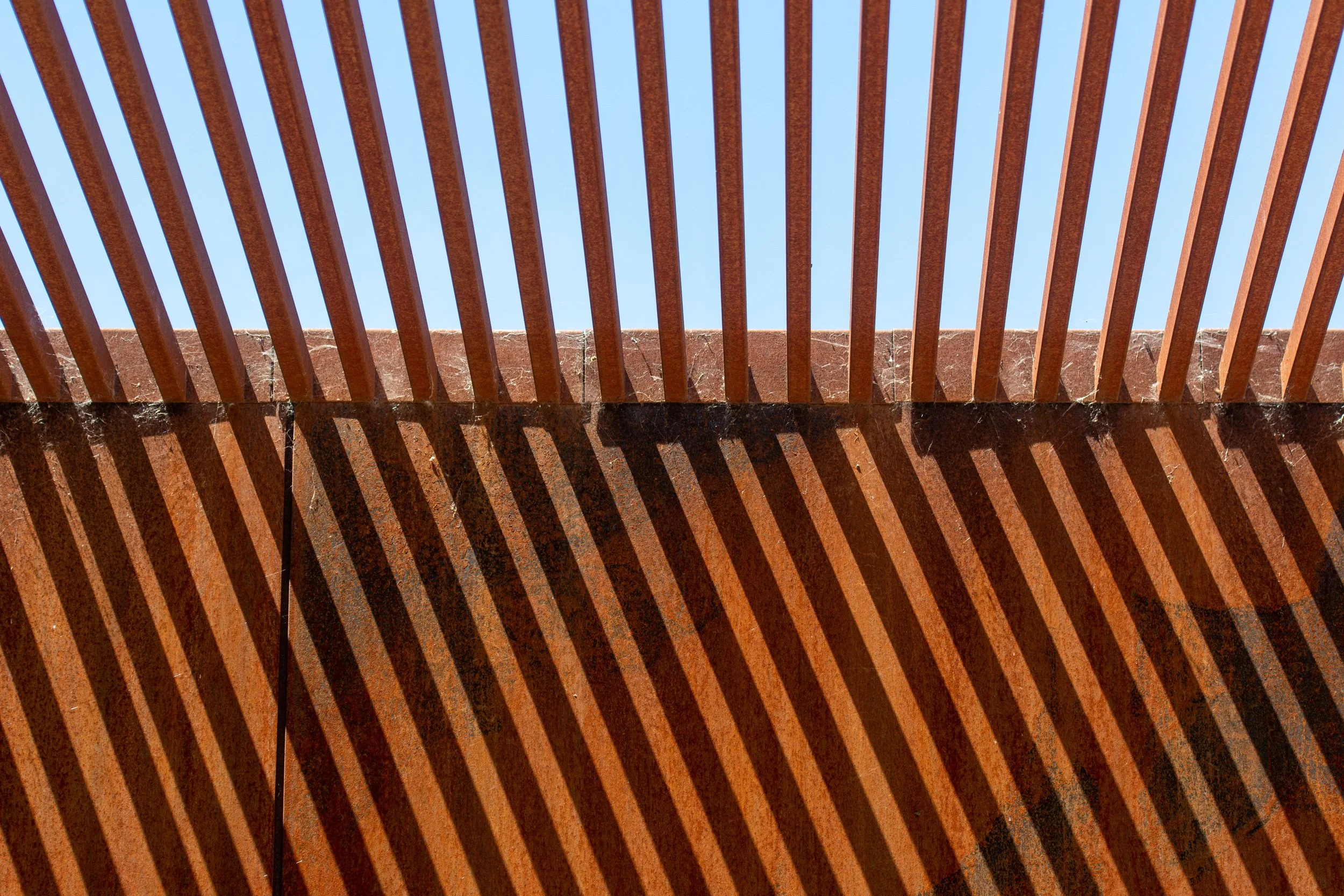 The photo shows a structure with horizontal reddish-brown wooden slats casting striped shadows against a bright blue sky.