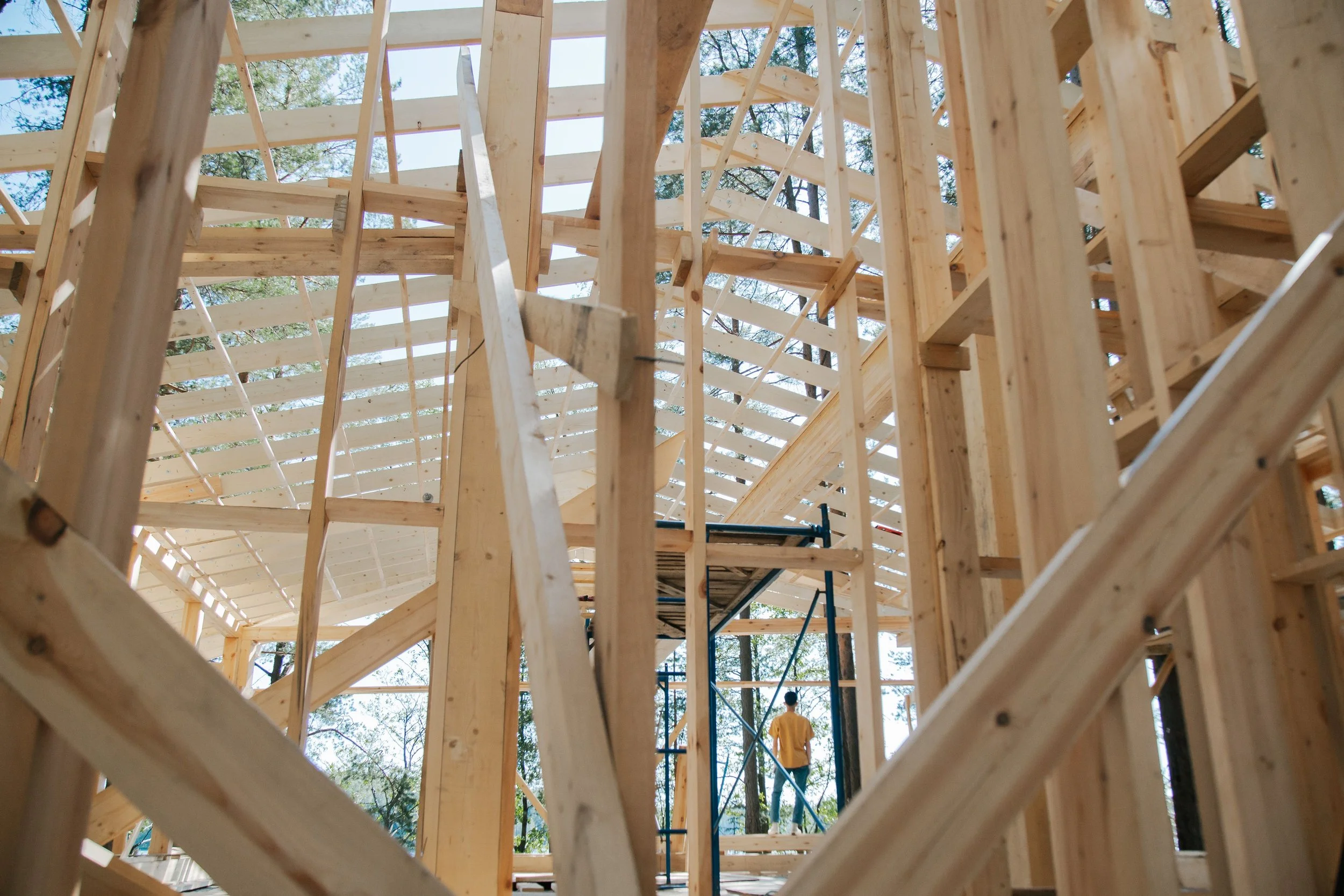 The image shows the wooden framing of a building under construction, with scaffolding and a worker in the background.