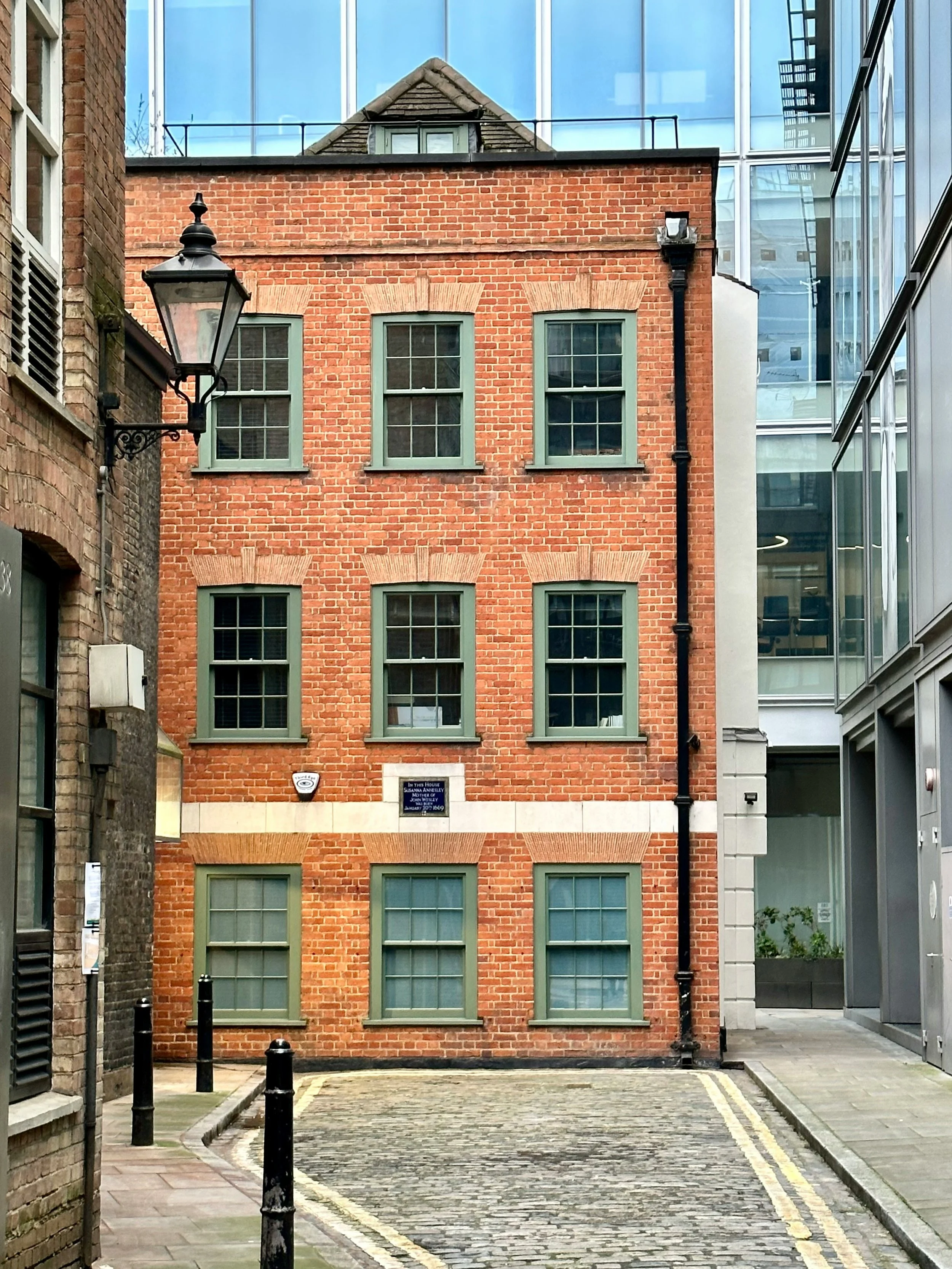 A narrow cobblestone alleyway bordered by brick and glass buildings, with a traditional street lamp and black bollards lining the sidewalk.